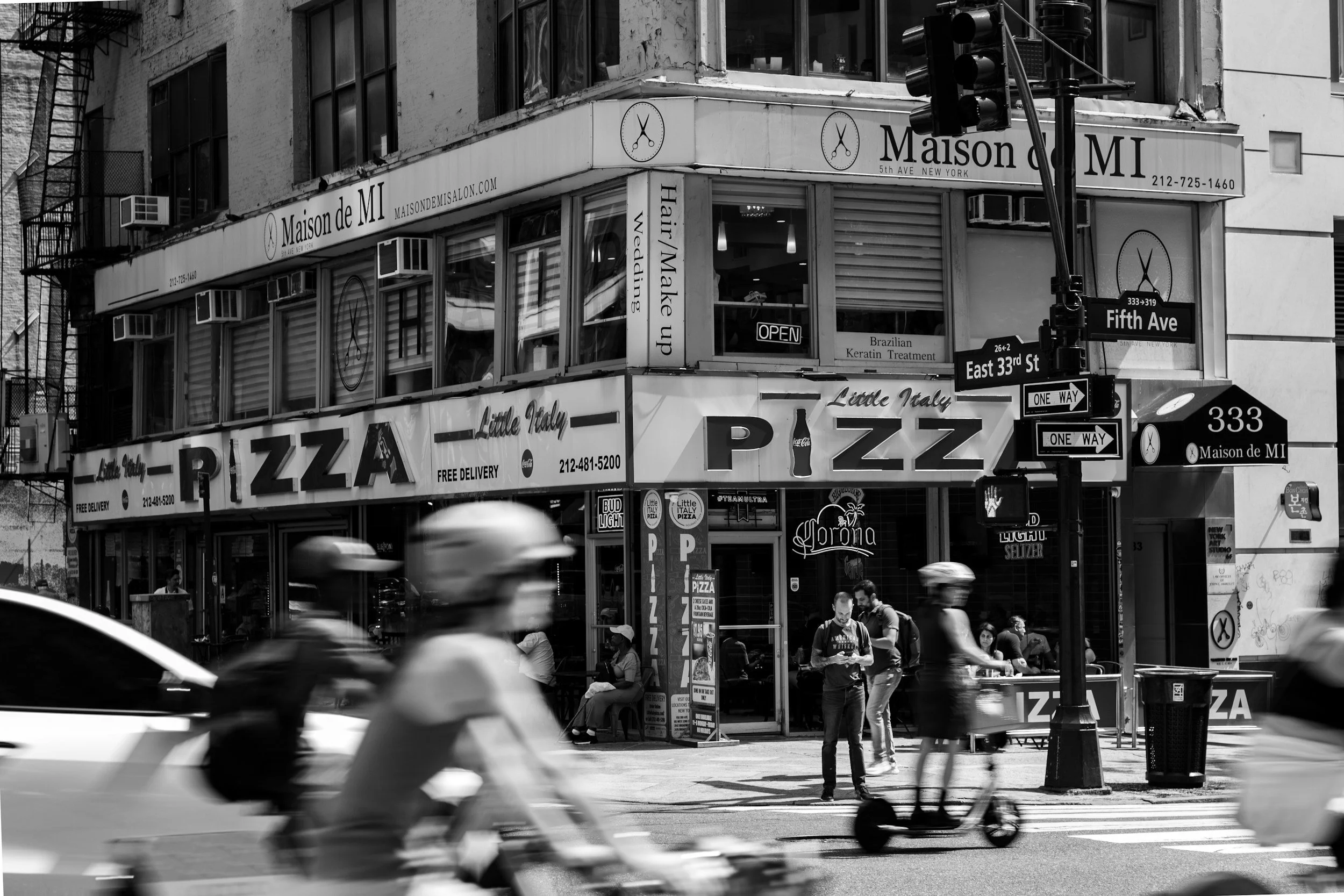 Black and white photo of a corner pizza restaurant called Little Italy on a busy city street, with pedestrians, bicycles, and a car passing by. Street signs for Fifth Avenue and East 33rd Street are visible.