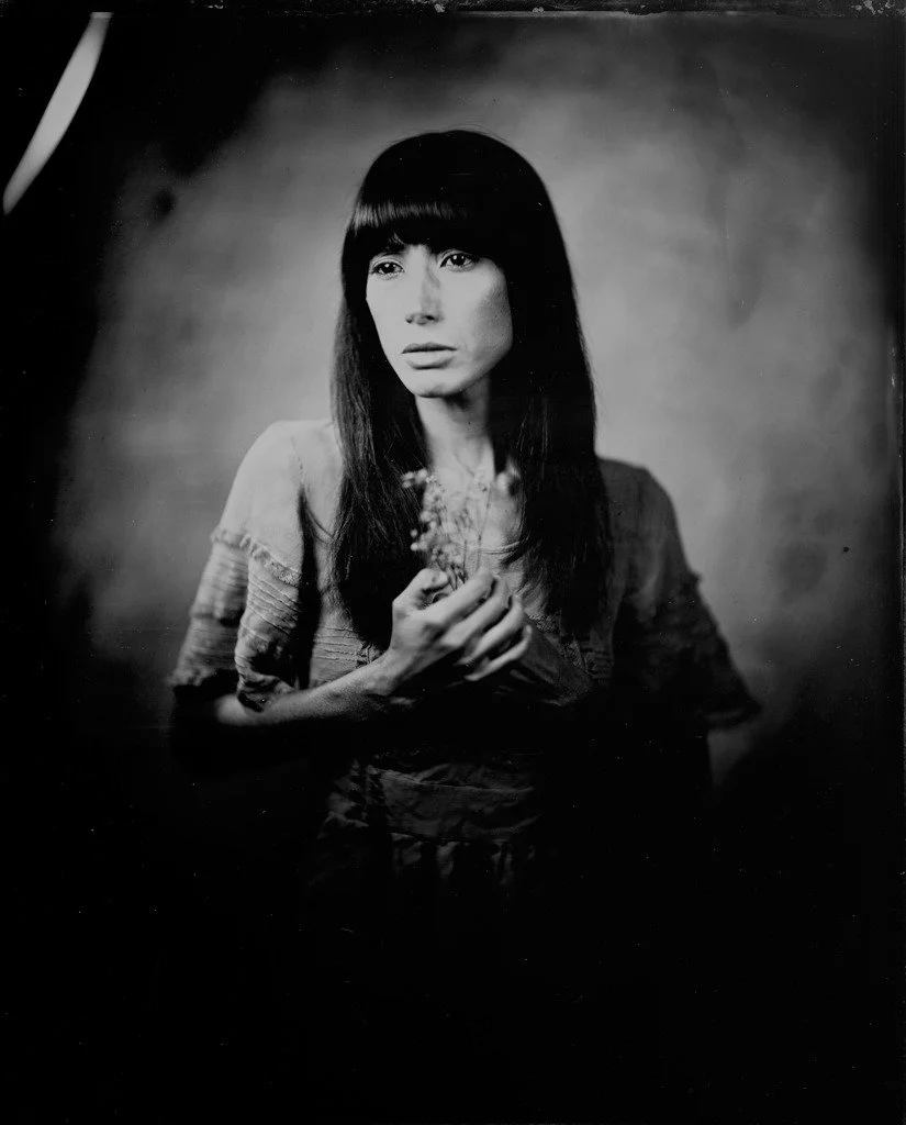 Black and white portrait of a woman with long, straight dark hair and bangs, holding a small plant or flower in her hands, looking slightly to the right.