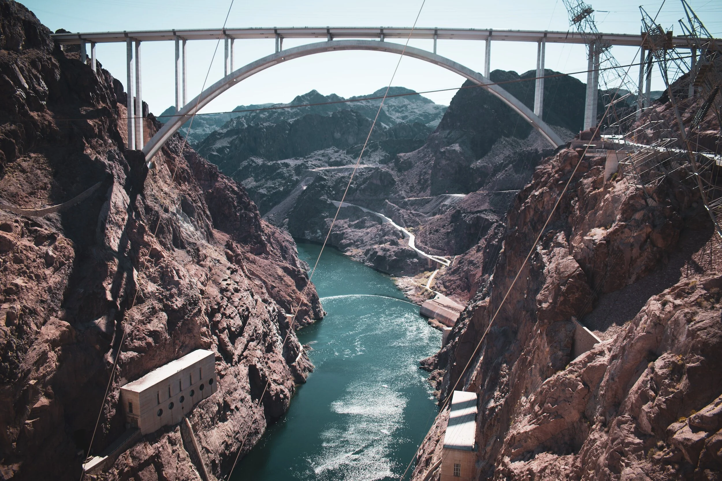 Image of the Hoover Dam with a large arch bridge high above the rocky canyon walls and the Colorado River flowing below.