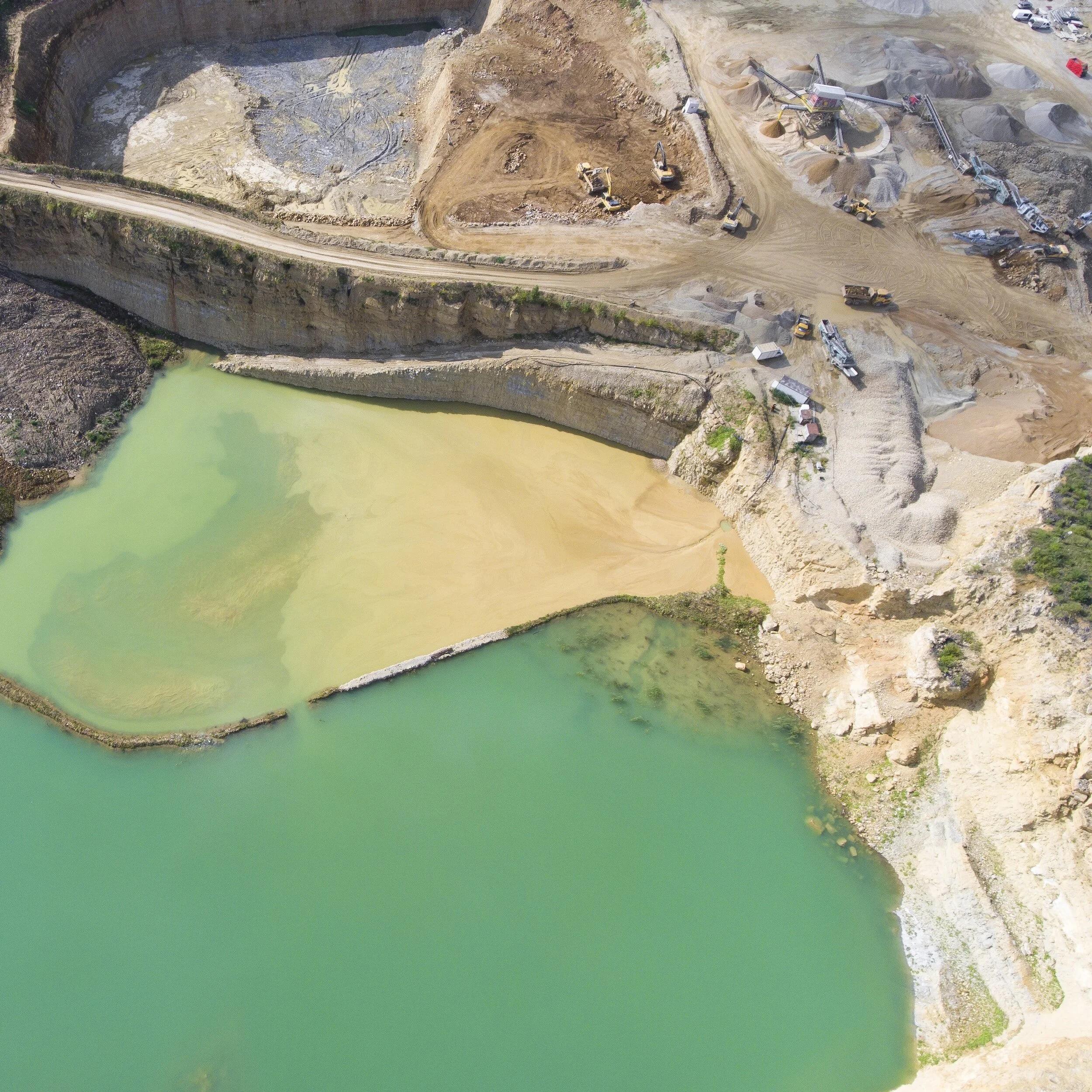Aerial view of a construction site near a large body of water with green and yellowish hues, showing excavators, trucks, and machinery on the land.