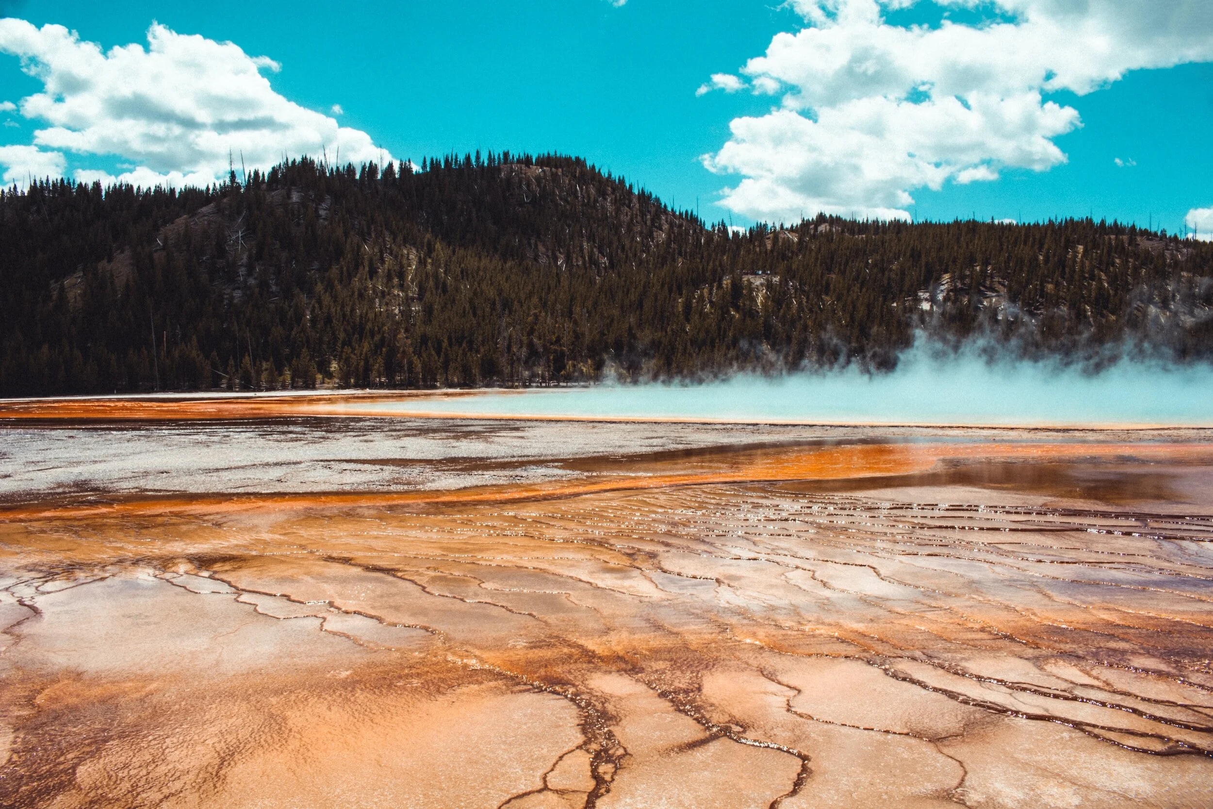 Colorful geothermal area with mineral deposits, steam rising, forested mountains, and a bright blue sky with clouds.