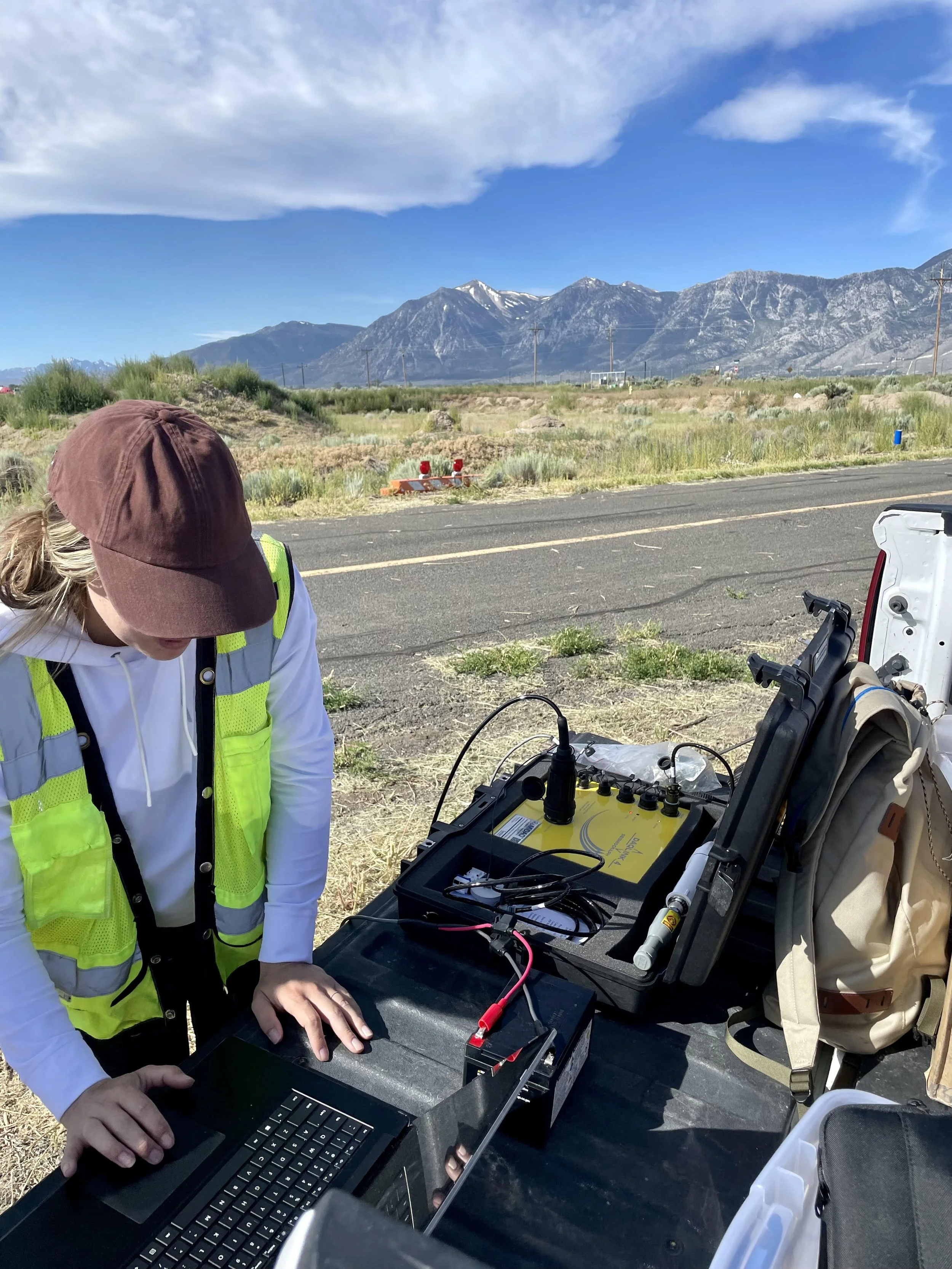 A person wearing a high-visibility vest and a brown cap is working on a laptop at a roadside setup with mountains in the background. Equipment including a yellow device, wires, and a backpack are on the table.