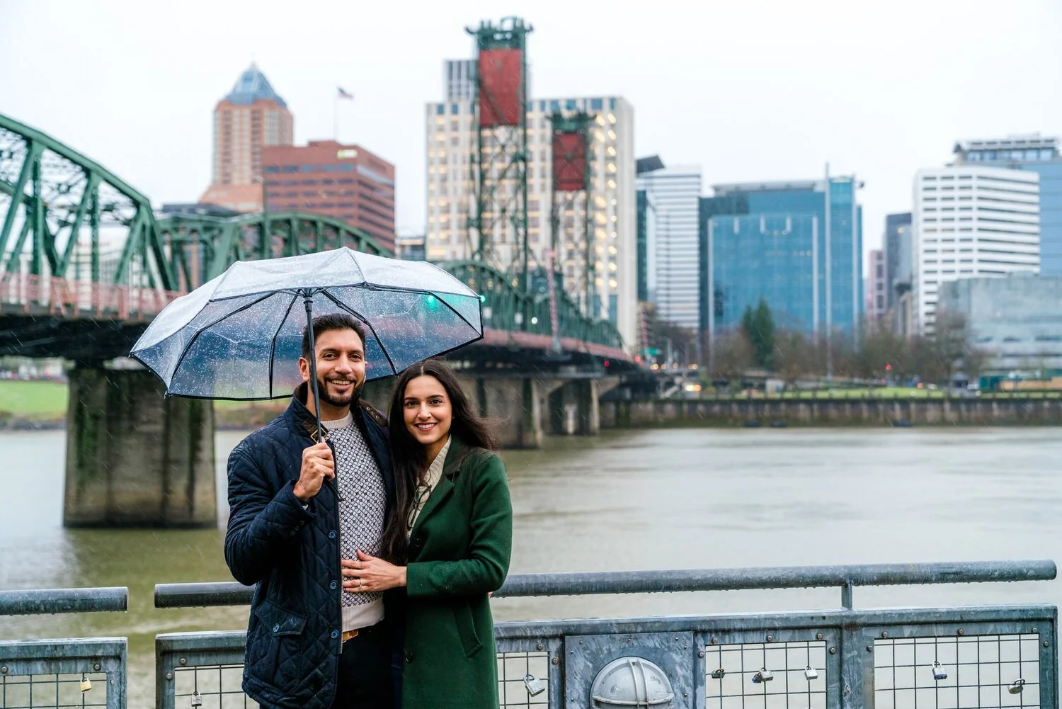Smiling couple standing under a clear umbrella on a rainy day, with a city skyline, river, and bridge in the background.