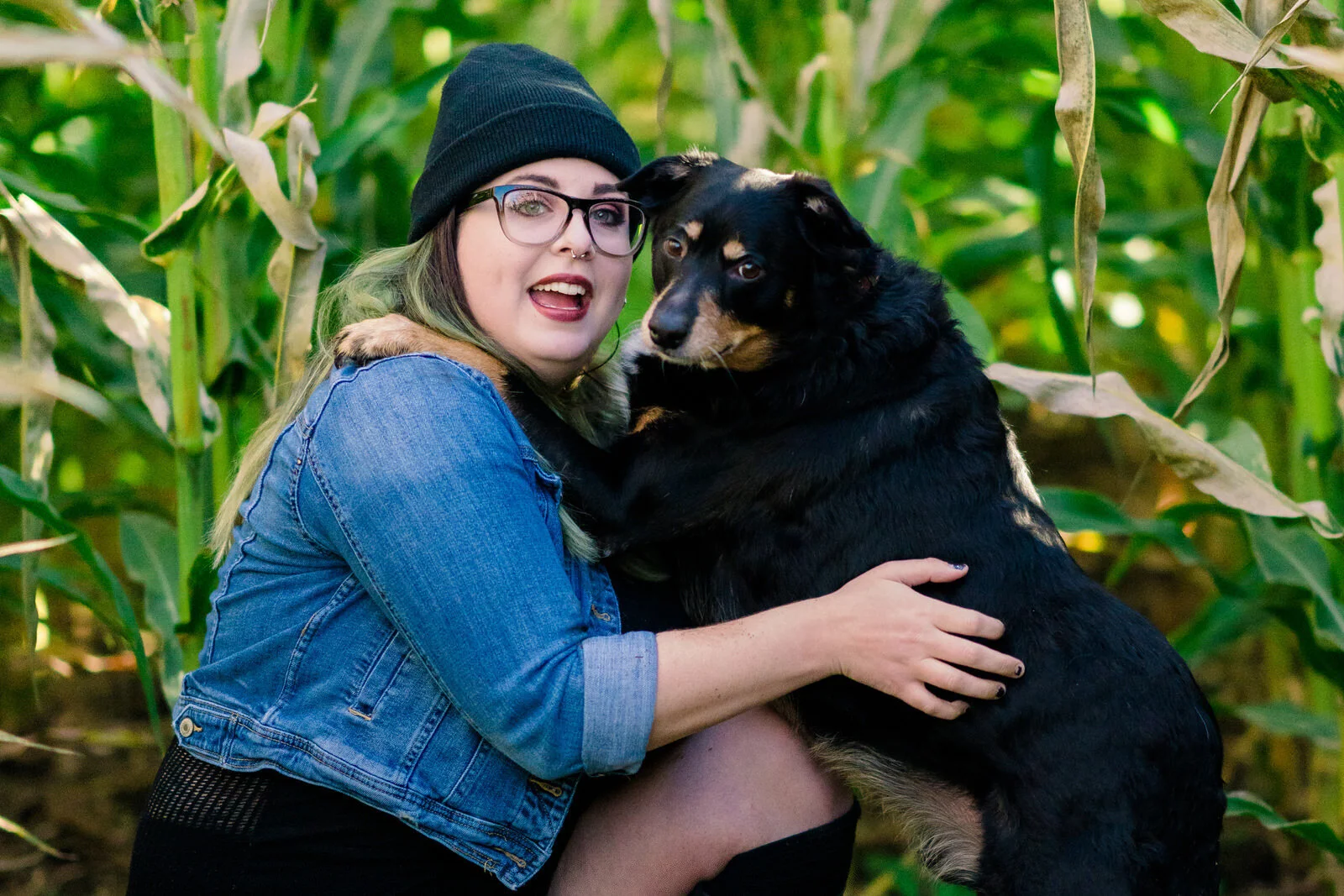 A woman in a denim jacket and black beanie smiles while hugging a black and tan dog in front of vibrant green cornstalks, showcasing a joyful moment outdoors.