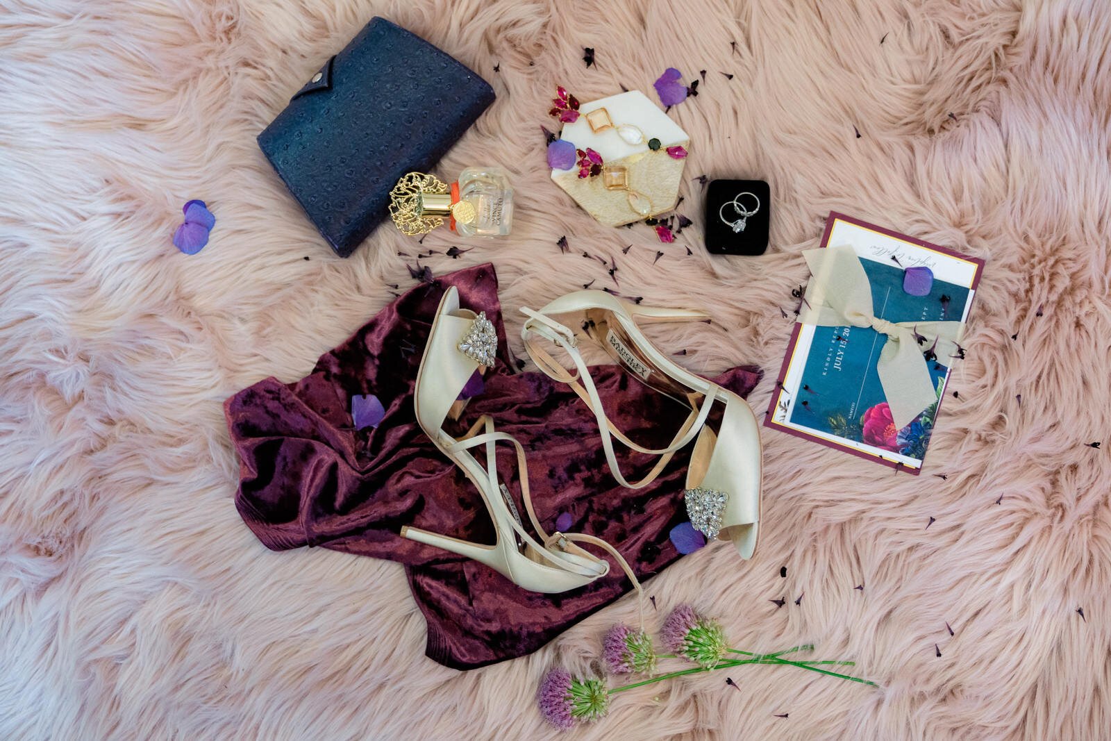 Elegant bridal flat lay with ivory heels, jewelry, perfume, a clutch, wedding invitations, and flowers arranged on a plush pink faux fur background with velvet fabric and petals.