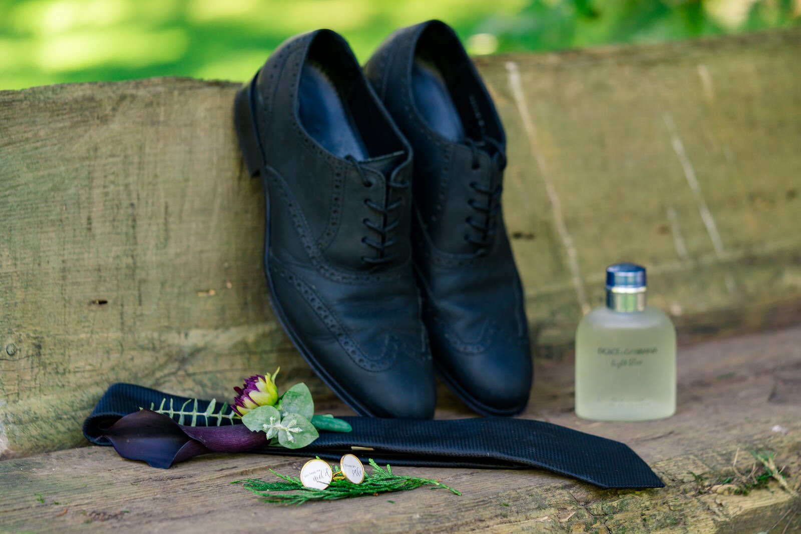 Groom’s wedding details including black dress shoes, a black tie, boutonniere, cufflinks, and cologne, arranged on a rustic wooden bench with a natural outdoor backdrop.