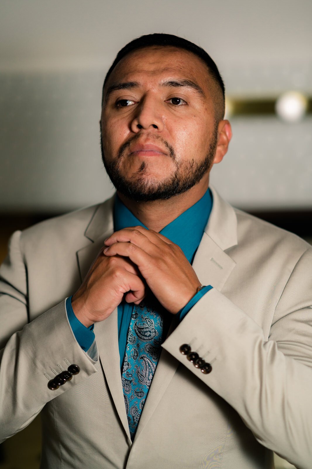 Groom in a beige suit and teal shirt adjusting his tie with a confident expression, preparing for his wedding day.