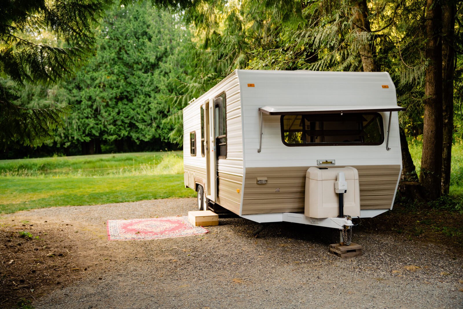 Vintage camper trailer parked in a scenic forest setting with a red patterned rug placed outside, creating a cozy and rustic atmosphere.