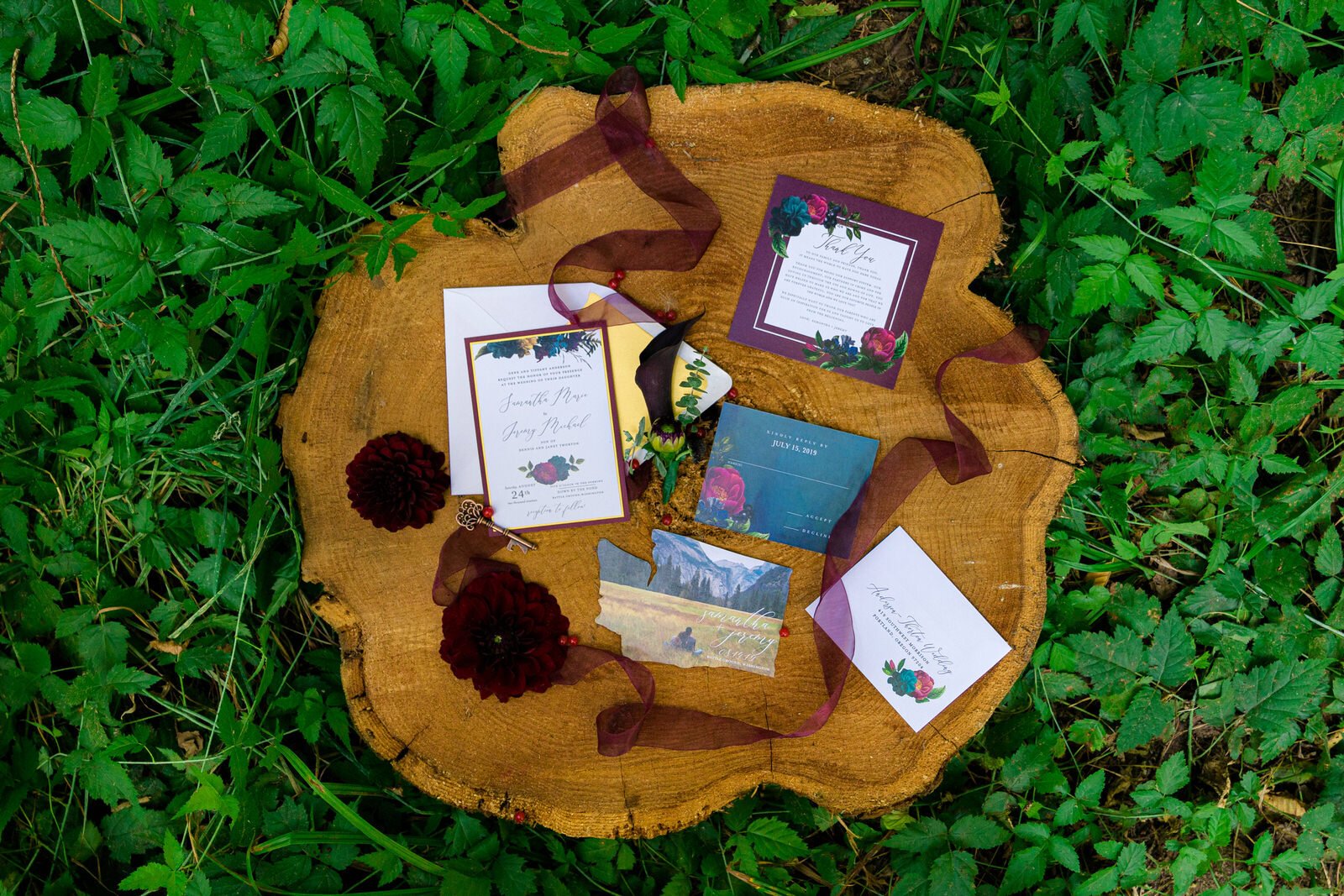 Elegant wedding invitation suite with floral details and deep jewel tones arranged on a rustic wood slab, surrounded by greenery and accented with ribbon and flowers.