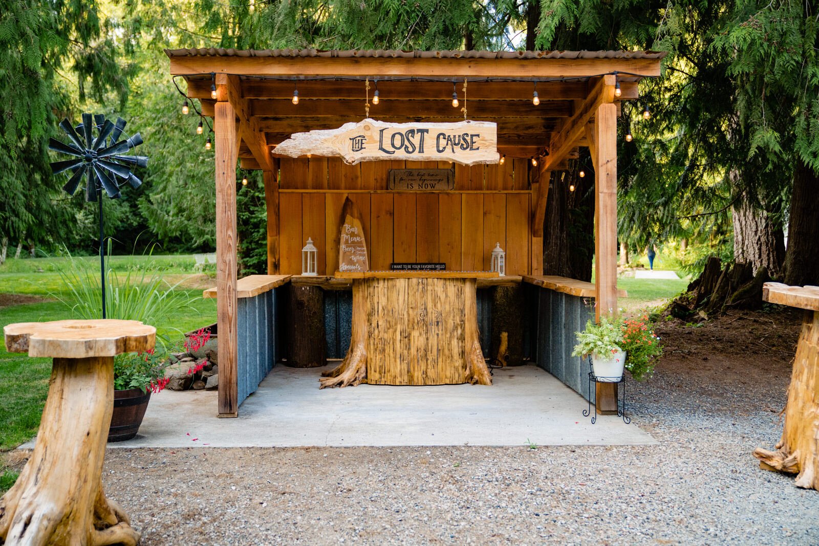 Rustic outdoor bar named “The Lost Cause,” built with wood and corrugated metal, adorned with string lights, surrounded by lush greenery and handcrafted wooden tables.