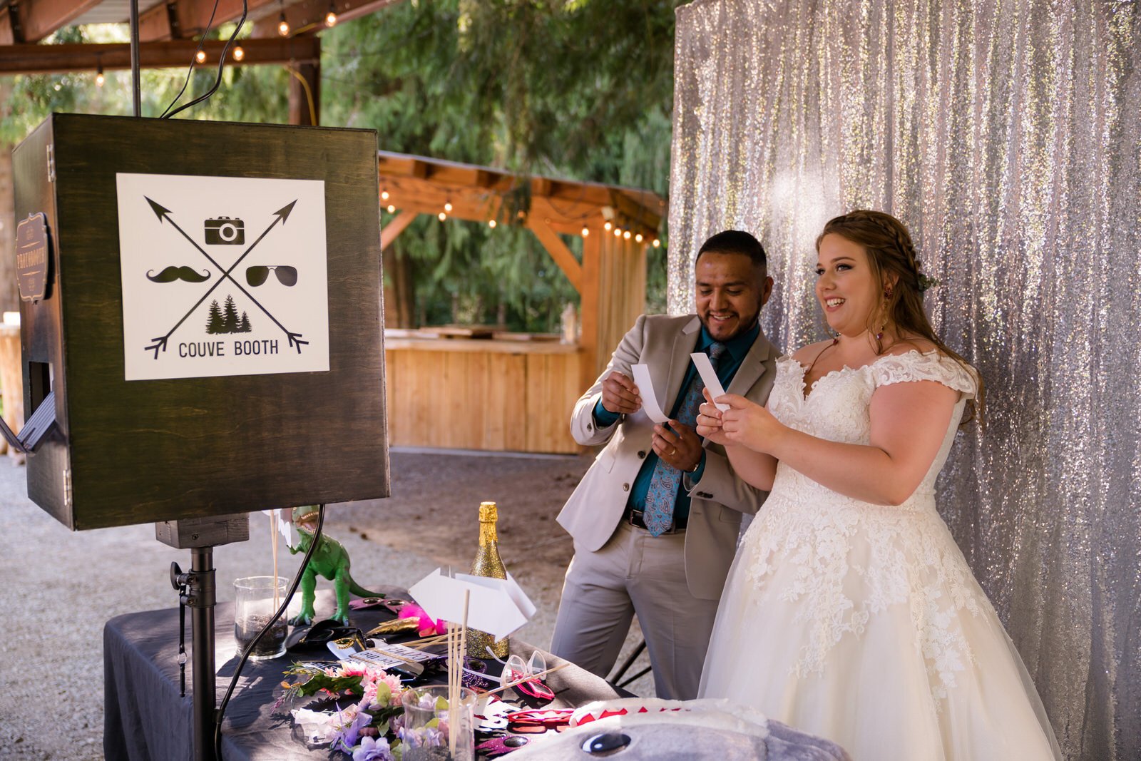 Bride and groom smiling as they look at photo booth prints at their wedding, with a rustic outdoor setting, silver backdrop, and fun props on the table.