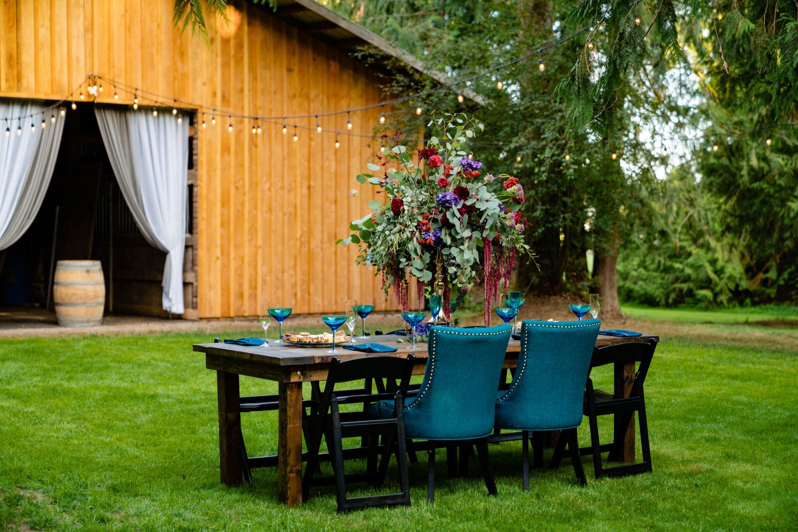 Elegant outdoor wedding reception setup with a rustic wooden table, teal chairs, vibrant floral centerpiece, and string lights, set against a barn with flowing white drapes.