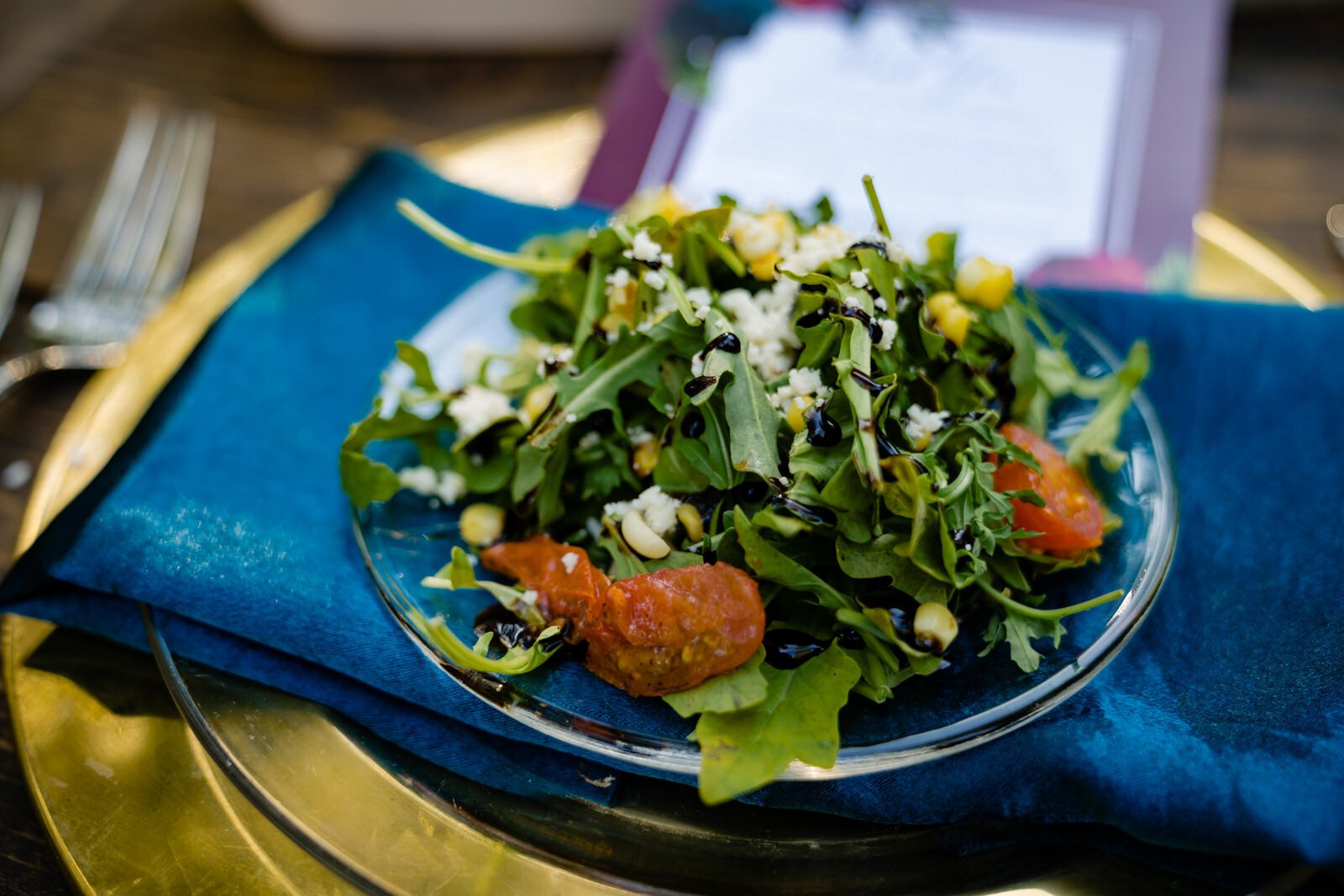 Fresh arugula salad with cherry tomatoes, corn, feta cheese, and balsamic drizzle, served on a glass plate over a blue napkin and gold charger at a wedding reception table setting.