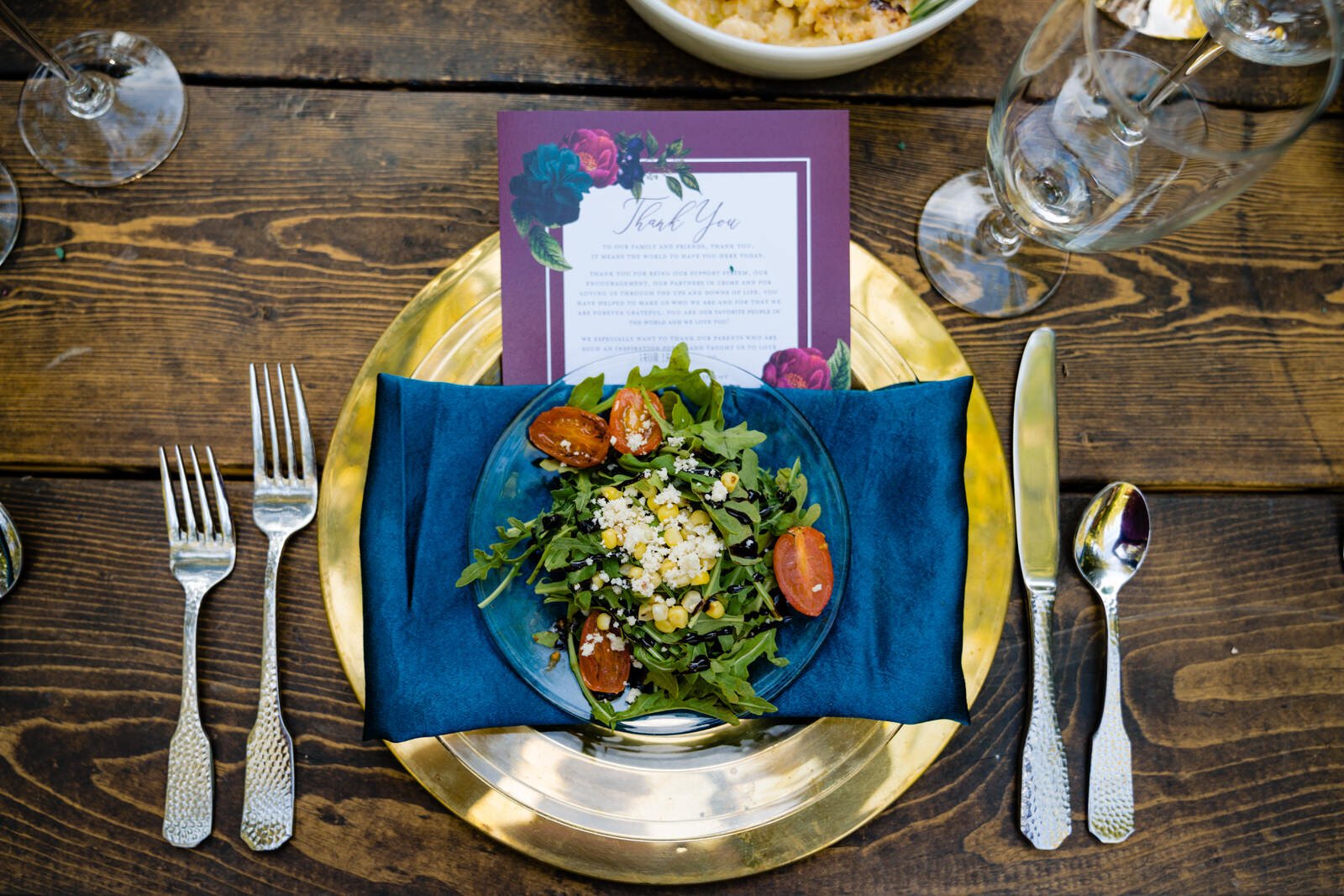 Elegant wedding table setting with a fresh arugula salad, gold charger, blue napkin, vintage silverware, and a floral thank-you card on a rustic wooden table with wine glasses.