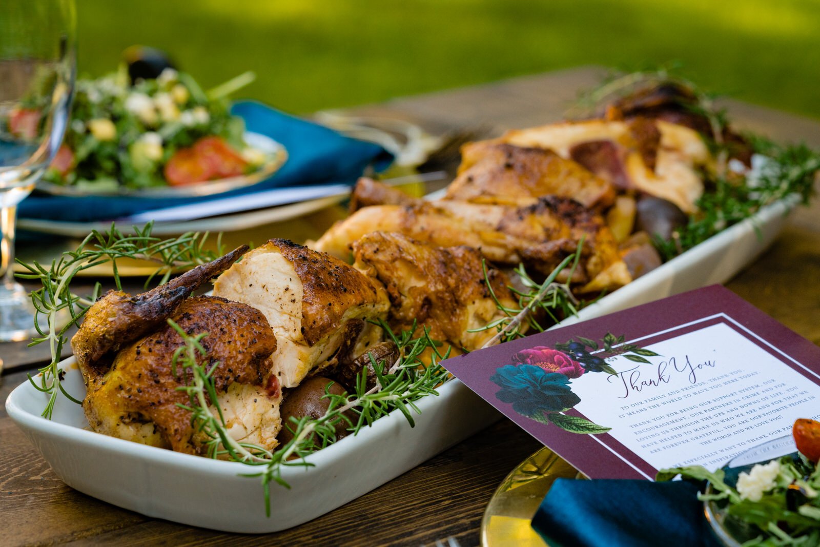Plated roasted chicken garnished with fresh rosemary at an elegant wedding reception table, with a thank-you card, salad, and wine glasses in the background.