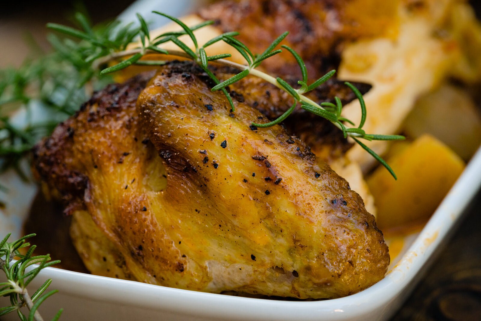 Close-up of golden, roasted chicken with crispy skin, garnished with fresh rosemary, served in a white dish with roasted potatoes.