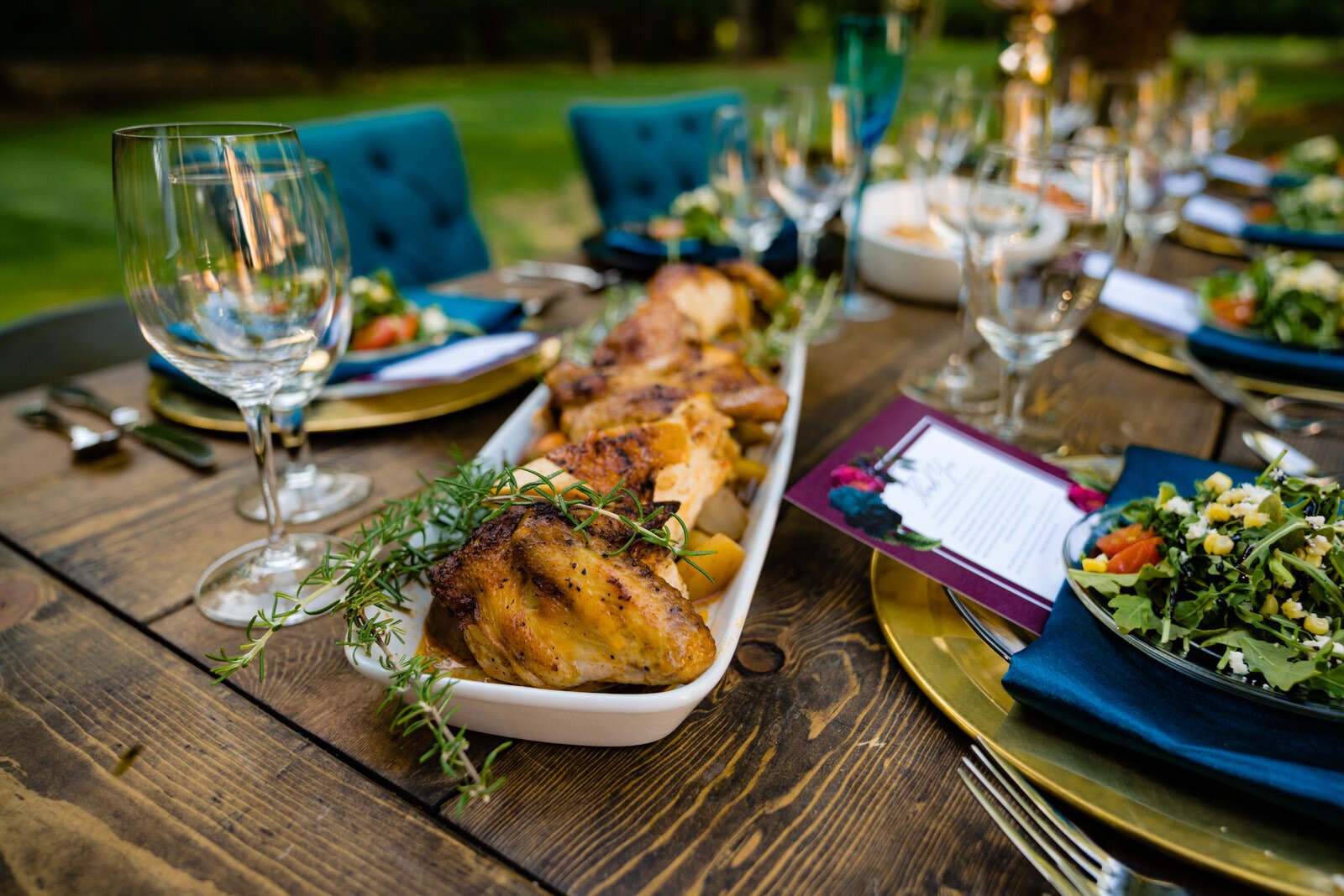 Elegant wedding reception table with plated roasted chicken, fresh salads, wine glasses, and blue napkins on a rustic wooden table with teal chairs and floral decor.