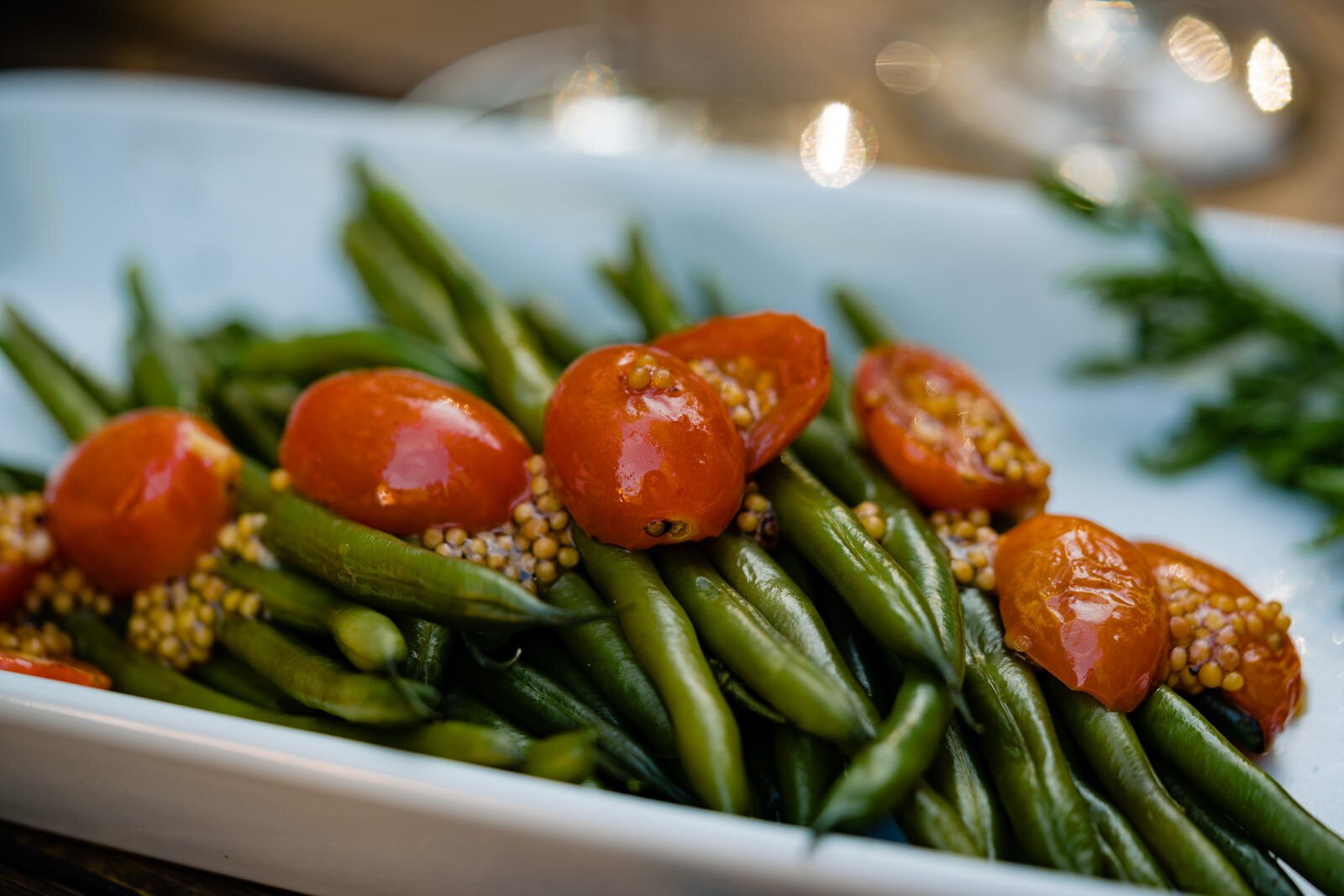Close-up of a plated side dish featuring green beans, roasted cherry tomatoes, and mustard seeds, served in a white dish at an elegant wedding reception.