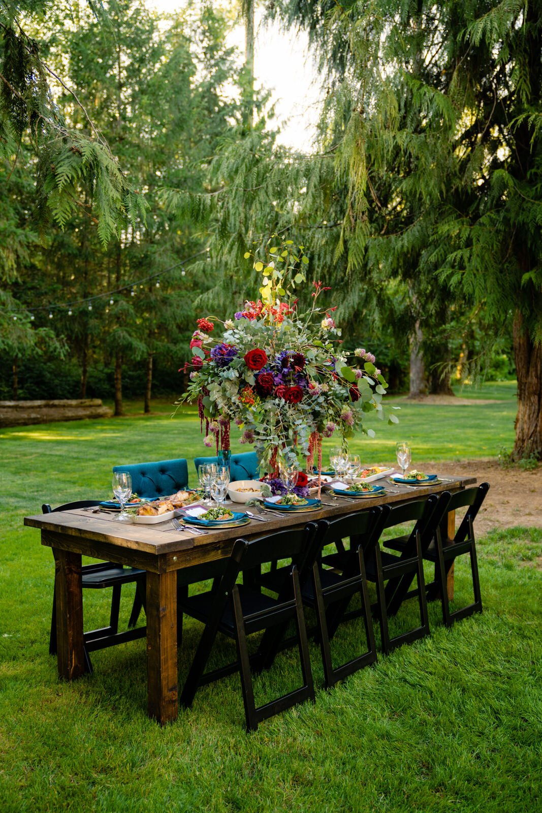 Outdoor wedding reception table with a vibrant floral centerpiece, elegant place settings, and rustic wooden decor, set in a lush green forest with string lights overhead.