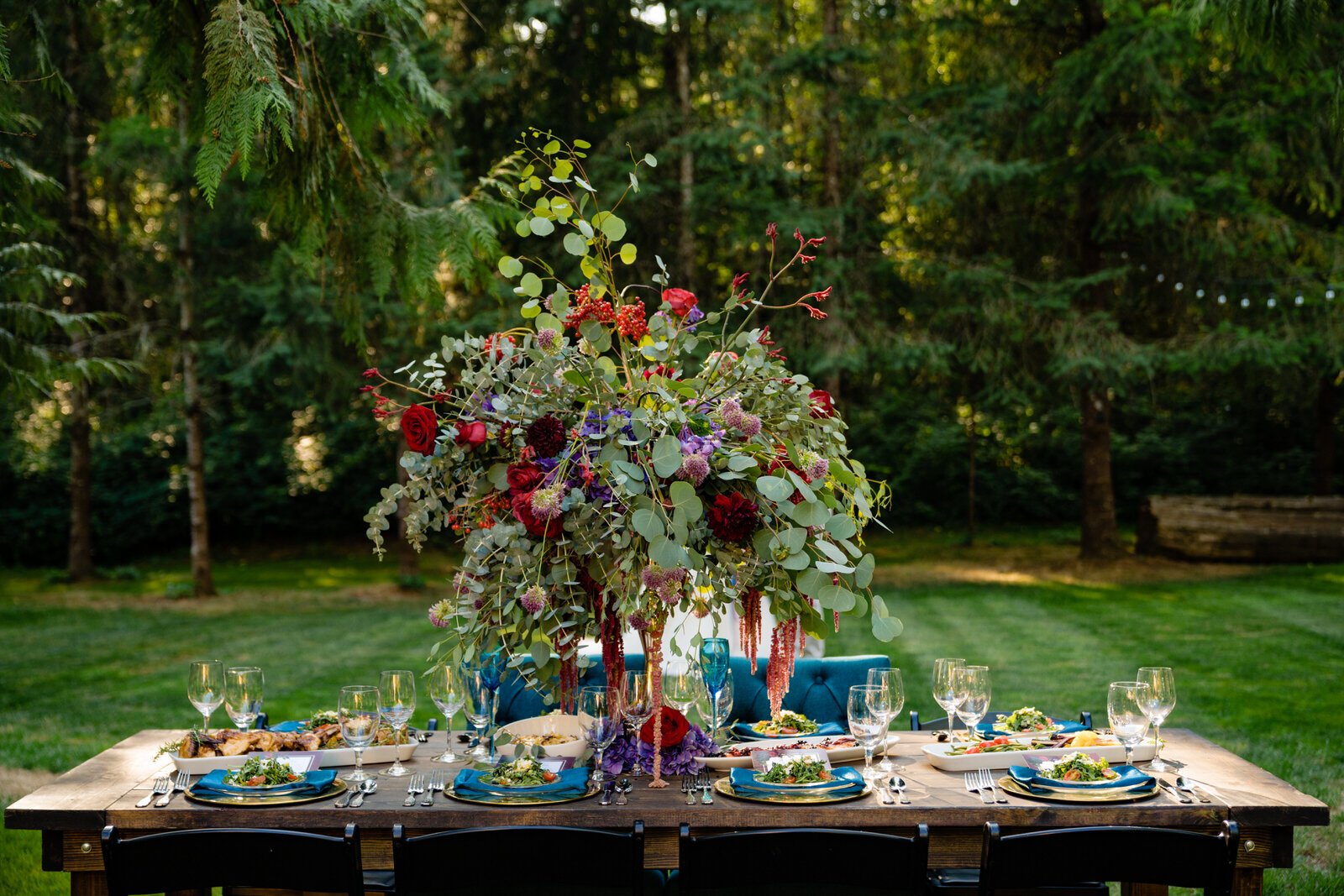 Outdoor wedding reception table with a lush floral centerpiece, elegant place settings, and gourmet food, set in a forested venue with natural greenery and string lights.