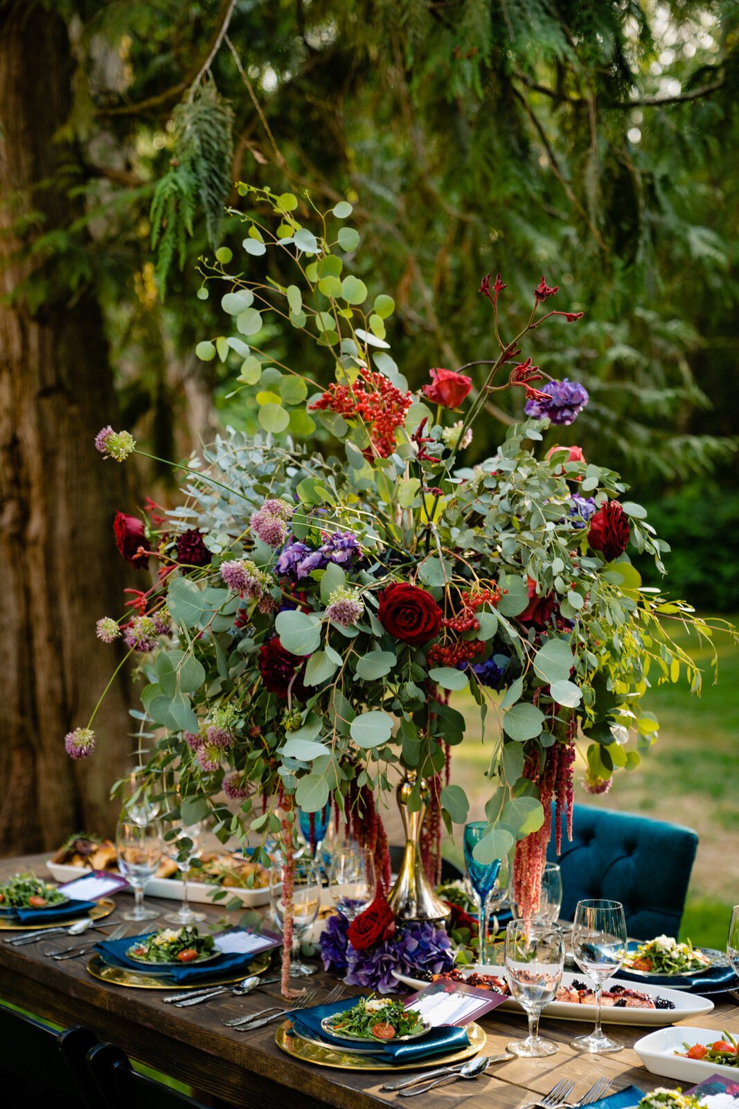 Lush wedding table centerpiece with deep red roses, eucalyptus, and cascading flowers, set on an elegantly decorated outdoor reception table with gourmet food and gold-accented place settings.