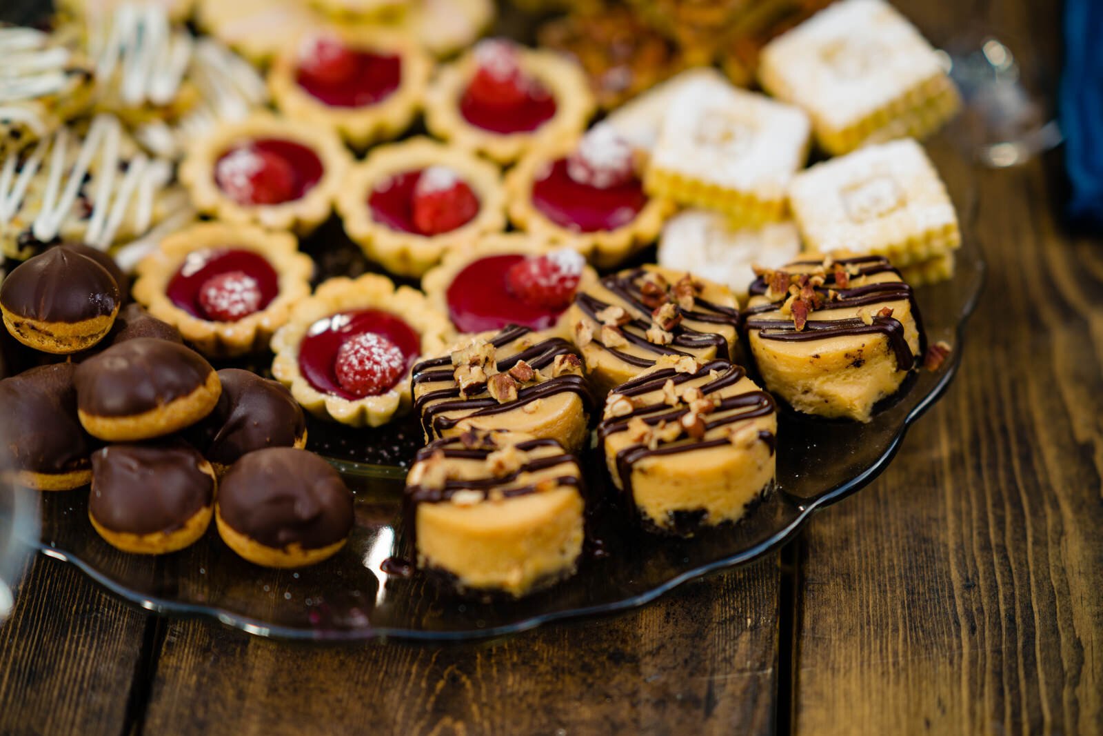 Elegant dessert platter featuring raspberry tarts, chocolate-drizzled mini cheesecakes with pecans, chocolate-covered cream puffs, and decorative cookies on a rustic wooden table.