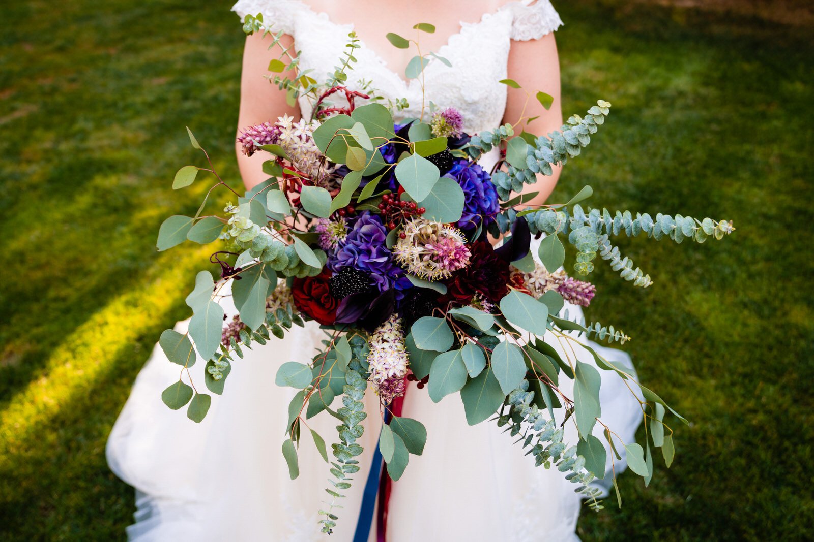 Bride in a white lace wedding dress holding a lush bouquet with eucalyptus, deep red and purple flowers, and wild greenery, set against a vibrant outdoor backdrop.