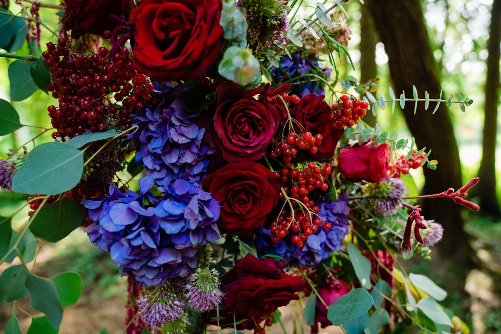 Close-up of a lush wedding floral arrangement featuring deep red roses, vibrant purple hydrangeas, red berries, and eucalyptus leaves, set against a forest backdrop.