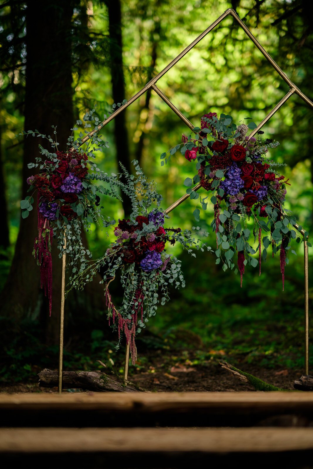 Modern geometric wedding arch adorned with deep red roses, purple hydrangeas, and eucalyptus, set in a lush forest ceremony space with rustic wooden benches.