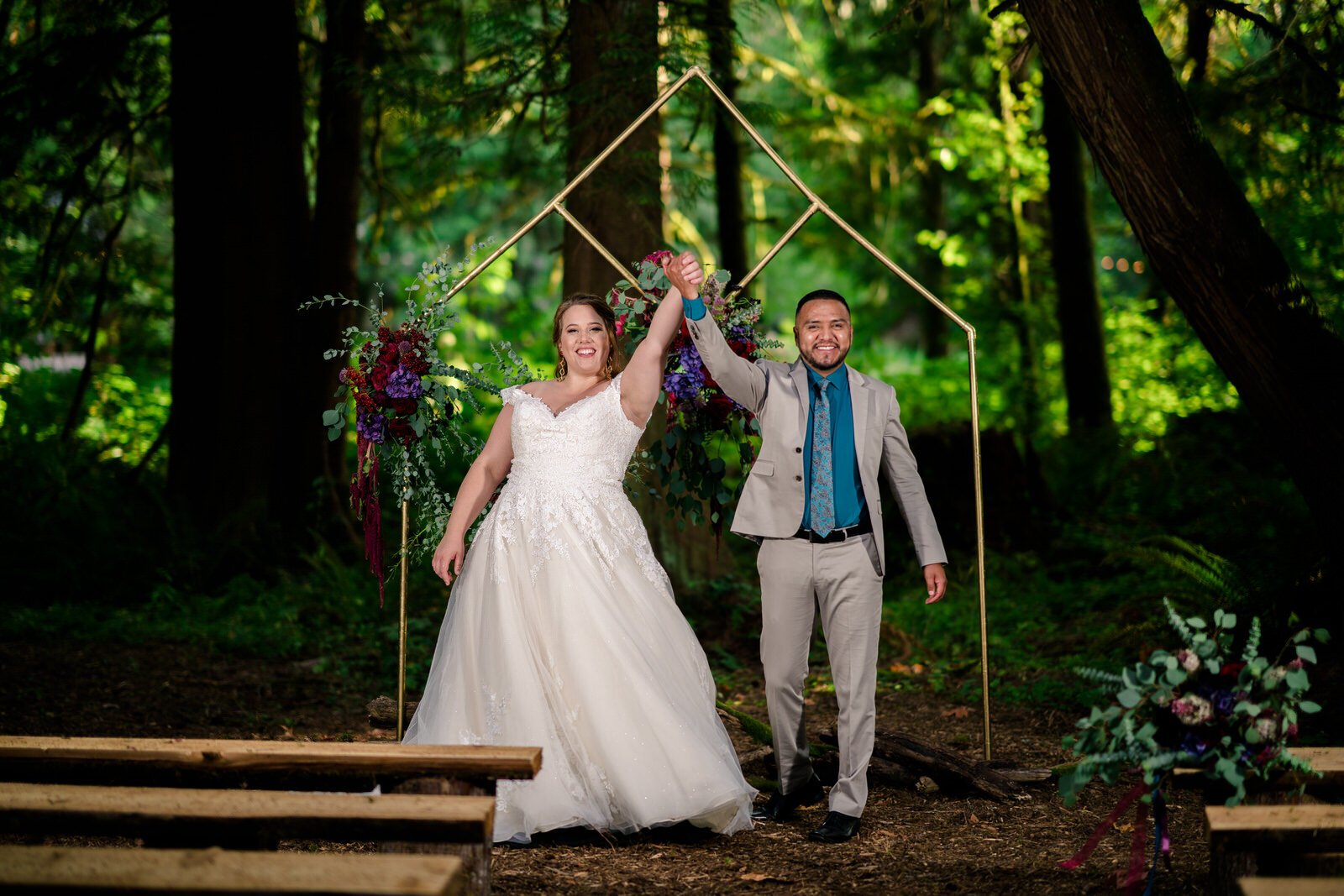 Newlywed couple celebrating under a modern geometric wedding arch adorned with flowers in a lush forest setting, holding hands joyfully after their ceremony.