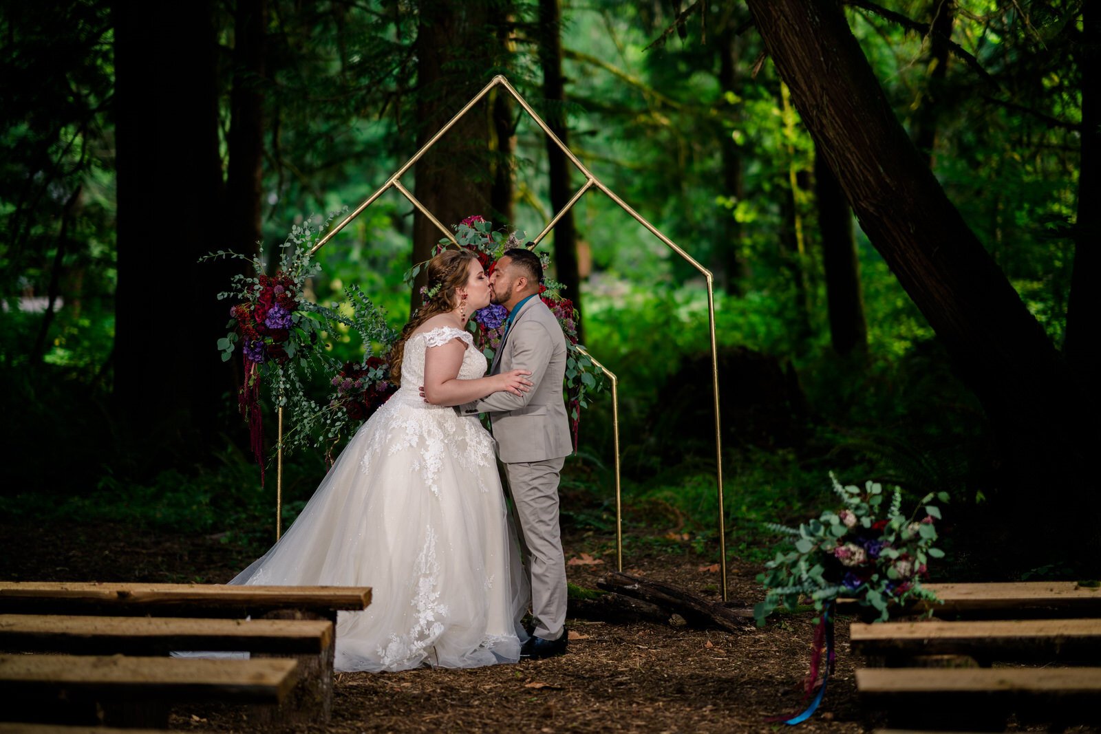 Bride and groom share a romantic kiss under a modern geometric wedding arch adorned with flowers in a lush forest ceremony setting with rustic wooden benches.