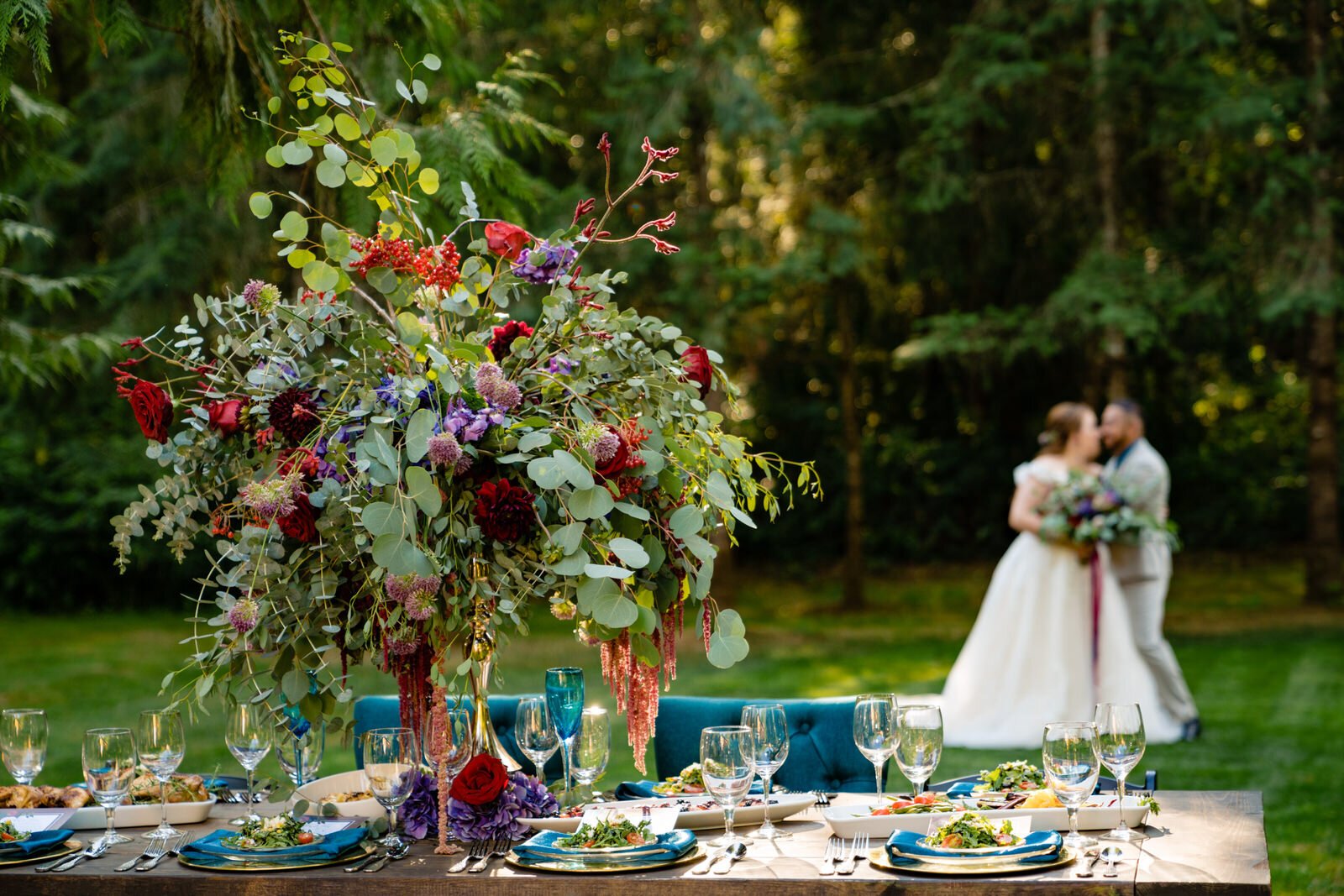 Elegant wedding reception table with a lush floral centerpiece, fine dining settings, and a blurred background of a newlywed couple embracing in a scenic forest.