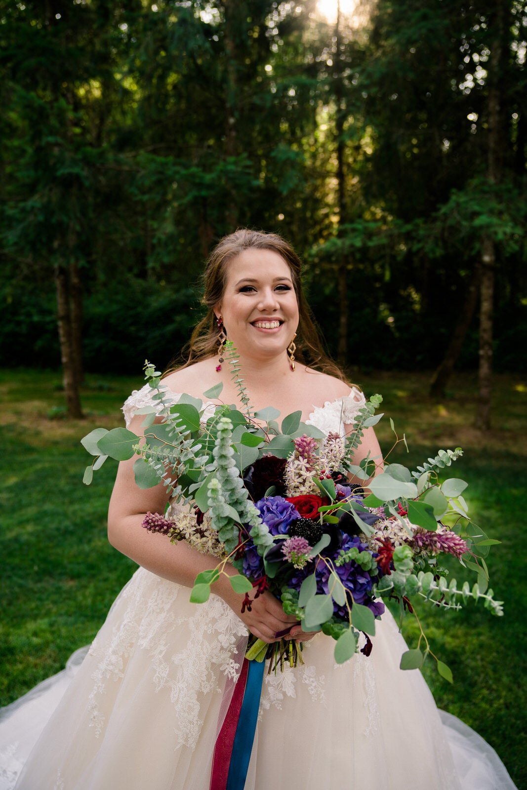 Smiling bride in a lace wedding dress holding a lush bouquet with eucalyptus, deep red and purple flowers, and ribbon accents, standing in a scenic forest setting.