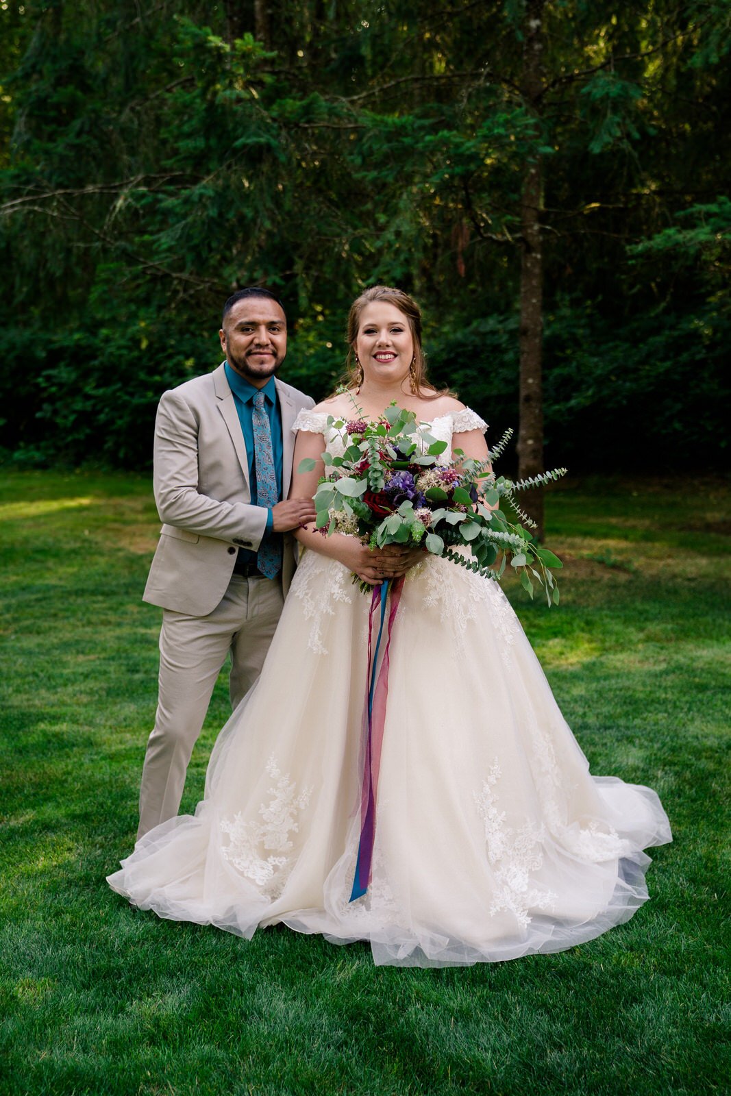 Bride in a lace wedding gown holding a colorful bouquet, standing beside the groom in a beige suit and blue tie, both smiling against a lush green forest backdrop.