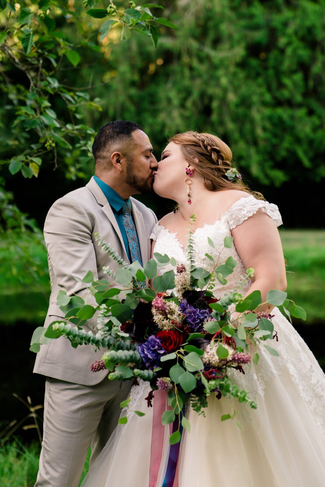 Bride in a lace wedding gown and groom in a beige suit share a kiss outdoors, with the bride holding a lush bouquet of red and purple flowers accented with eucalyptus greenery.