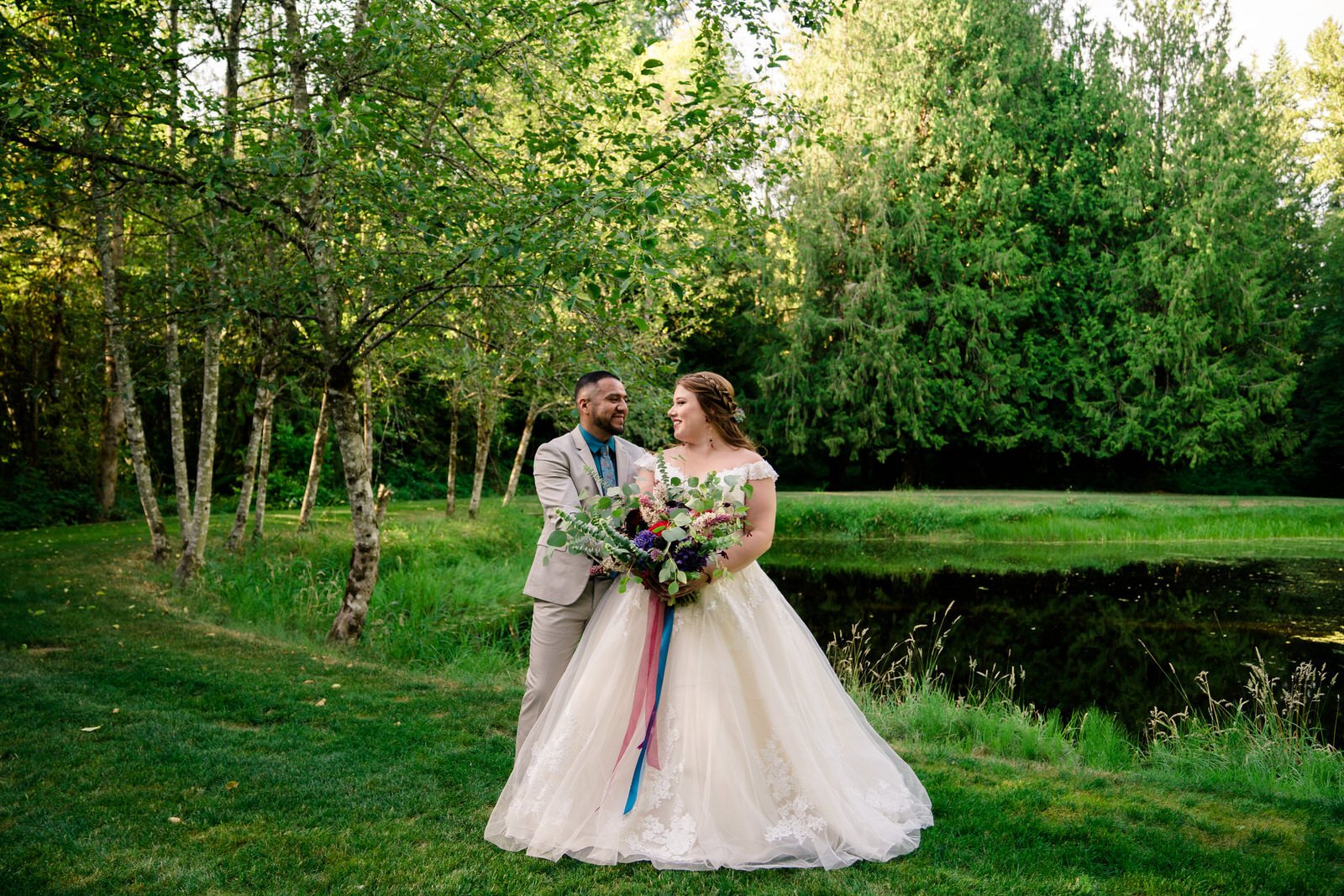 Bride in a flowing lace wedding gown and groom in a beige suit stand together by a reflective pond, surrounded by lush greenery and trees, sharing a joyful moment on their wedding day.