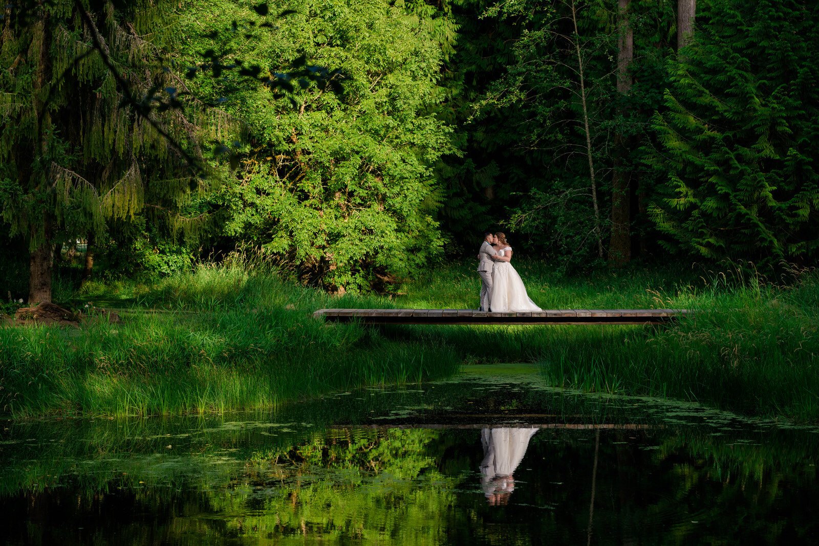 Bride and groom embrace on a wooden dock surrounded by lush greenery, their reflection visible in the still pond water, creating a serene and romantic wedding scene in nature.