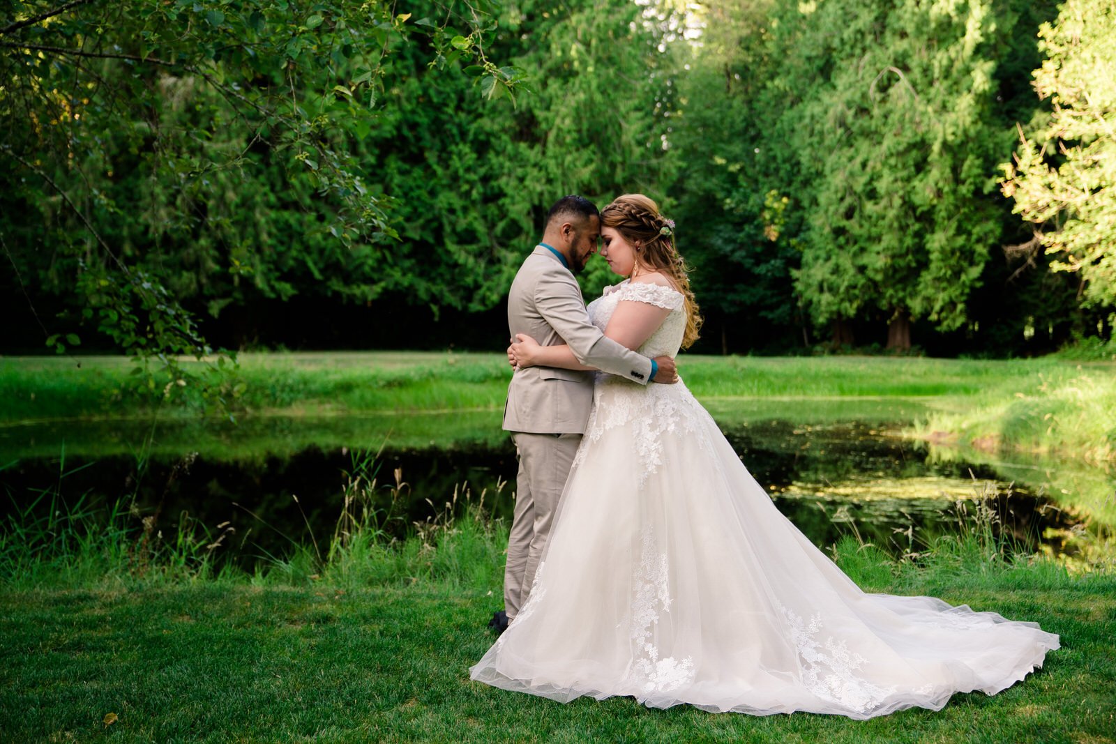 Bride and groom share a quiet, intimate moment by a serene pond, surrounded by lush greenery, during their outdoor wedding.