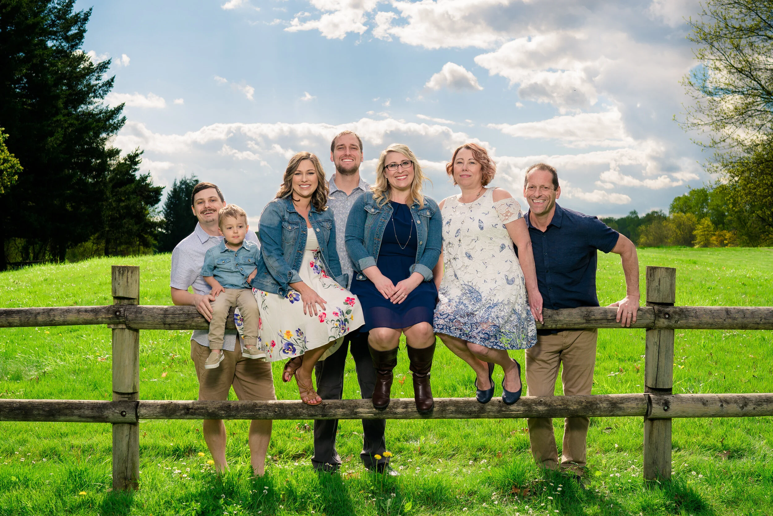 Family posing together outdoors on a wooden fence, with a green field, trees, and a cloudy sky in the background.