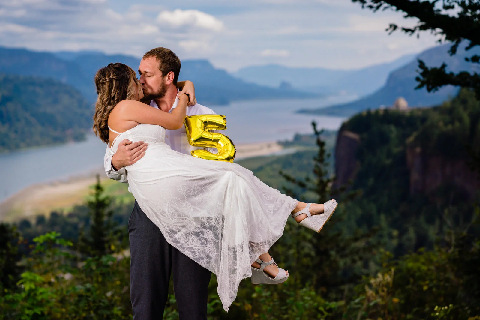 Couple celebrating their anniversary with a scenic kiss, holding a gold ‘5’ balloon, overlooking a breathtaking river and mountain landscape.
