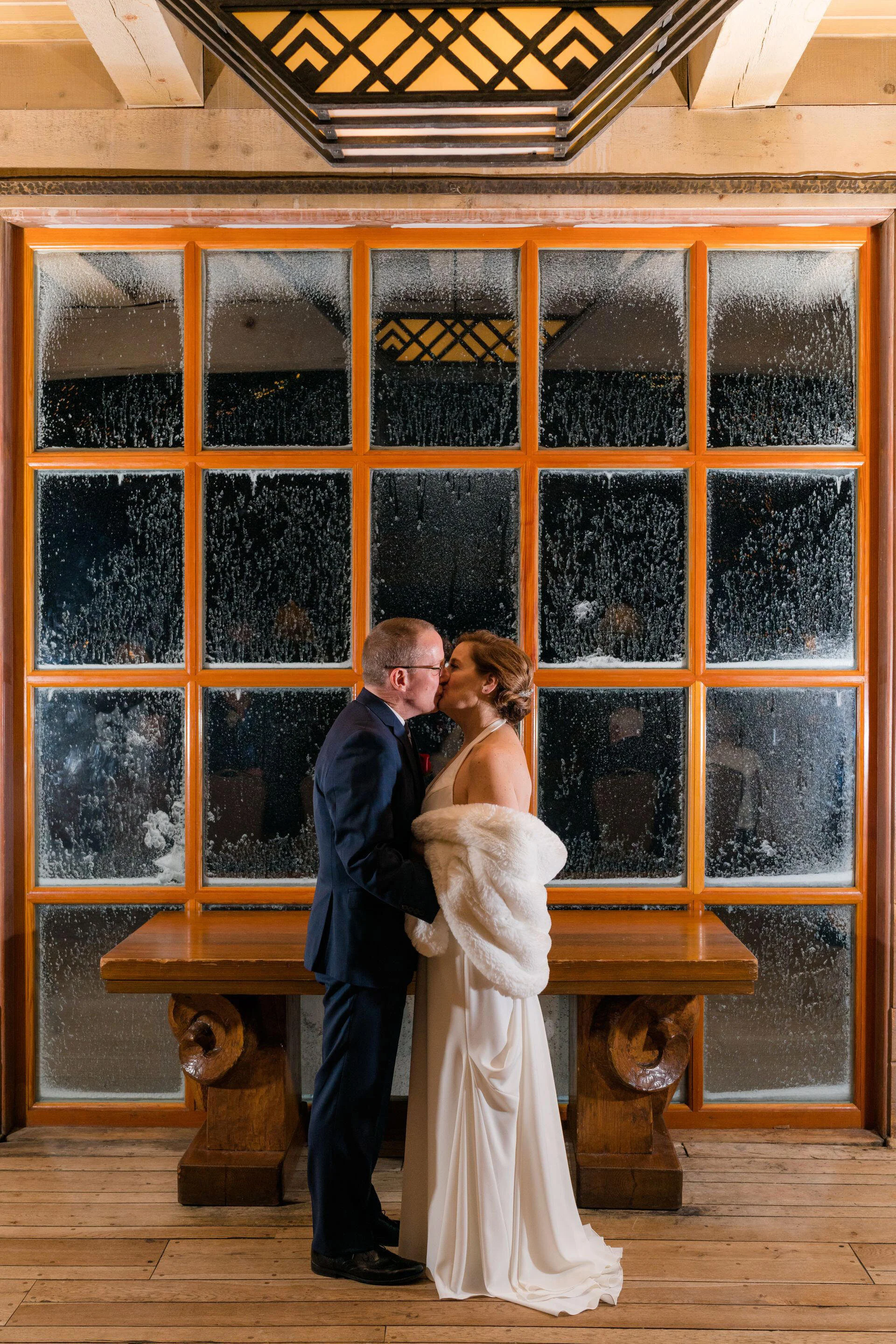 Bride and groom sharing a kiss indoors, framed by a large frosted window and warm wooden accents, with the bride wrapped in a white fur stole.