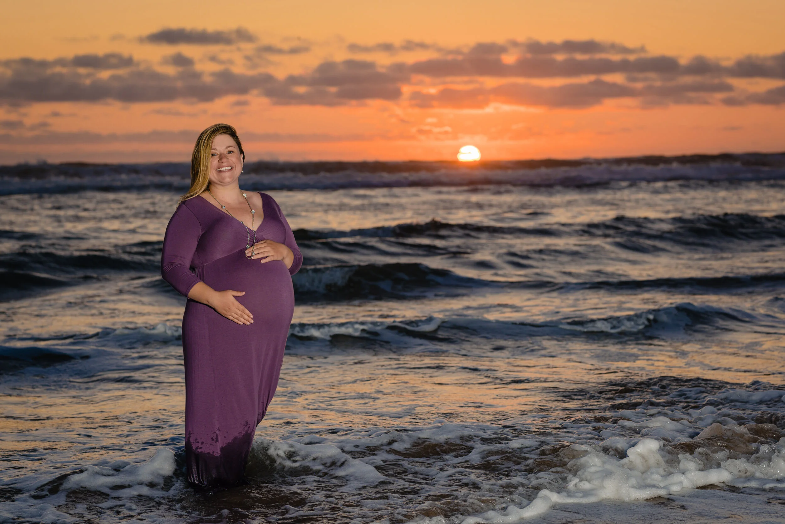 A pregnant woman in a long purple dress stands in the shallow ocean waves at sunset, smiling as the sun sets behind her over the water.