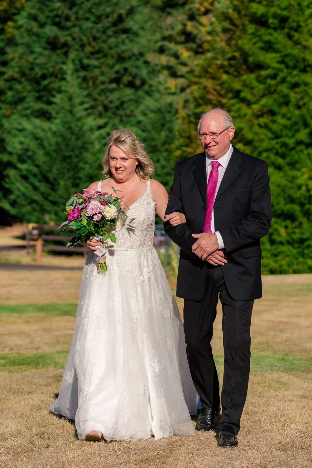 Bride walking down aisle with father at outdoor backyard wedding ceremony in Vancouver Washington, holding colorful bouquet.