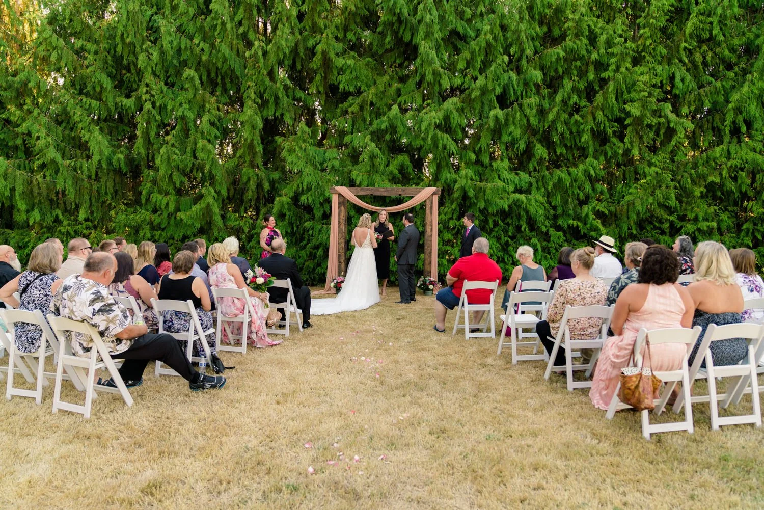 Wide view of intimate backyard wedding ceremony in Vancouver WA with wooden arch, blush draping, and guests seated on white chairs.