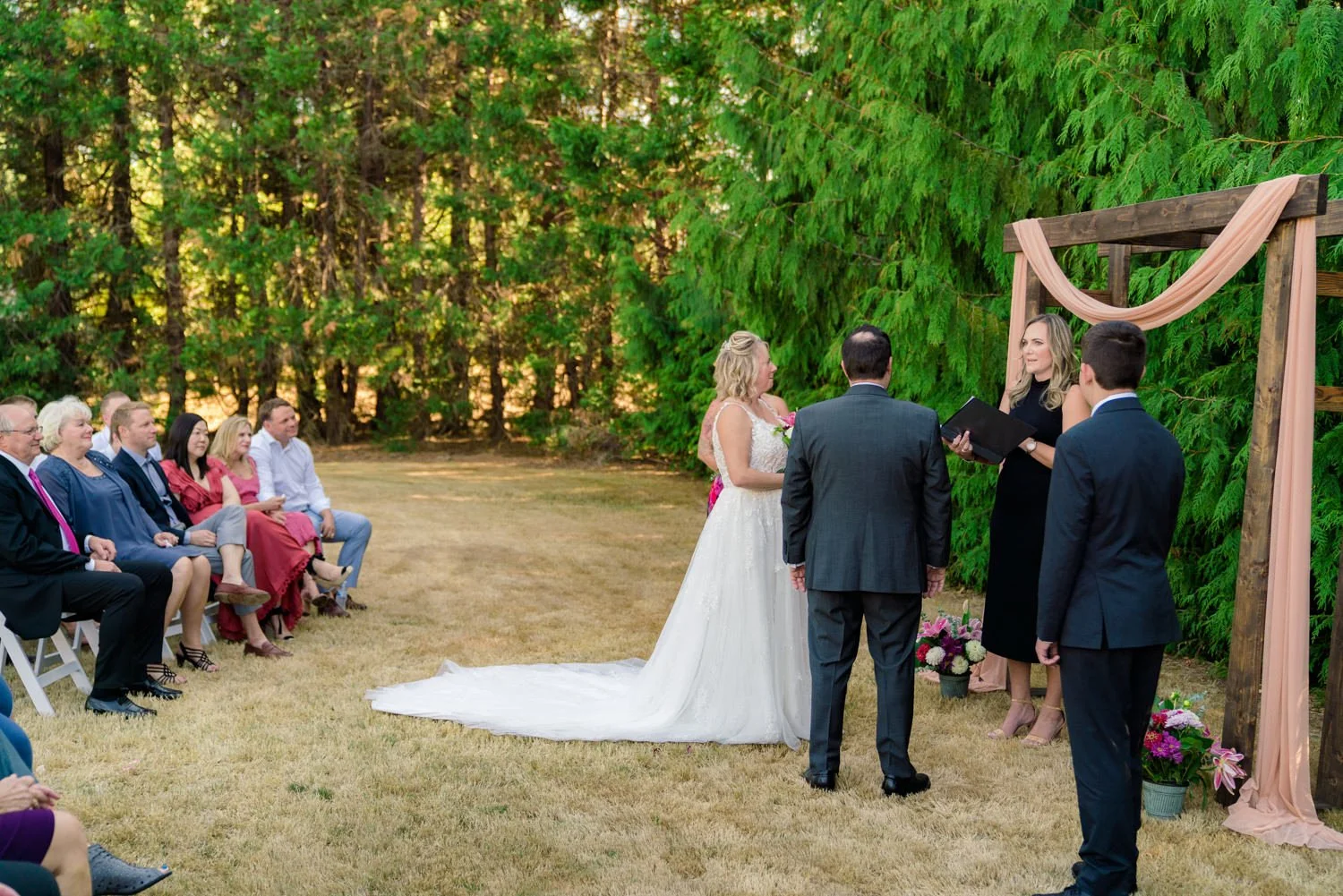 Bride and groom standing at wooden arch during backyard wedding ceremony as officiant speaks, surrounded by greenery in Vancouver WA.