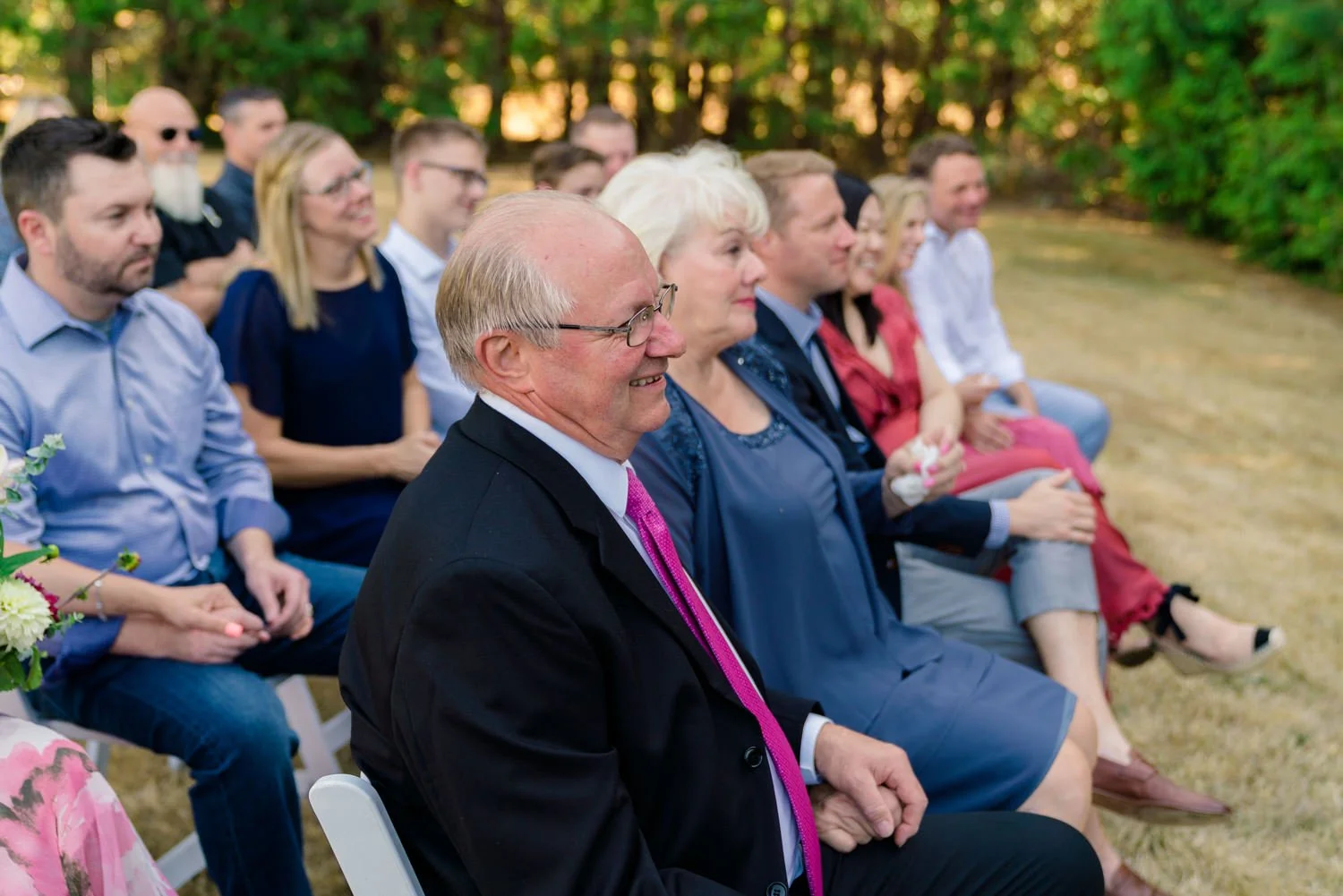 Wedding guests smiling and watching ceremony at intimate backyard wedding in Vancouver Washington with evergreen tree backdrop.