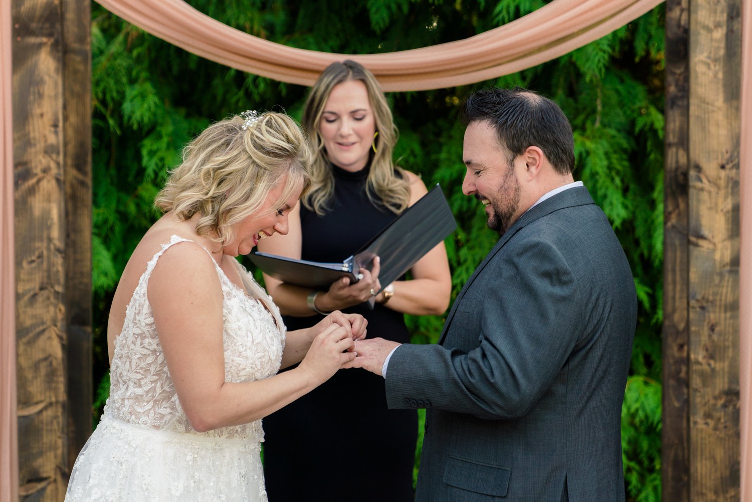 Bride and groom laughing during ring exchange at elegant backyard wedding ceremony under wooden arbor.
