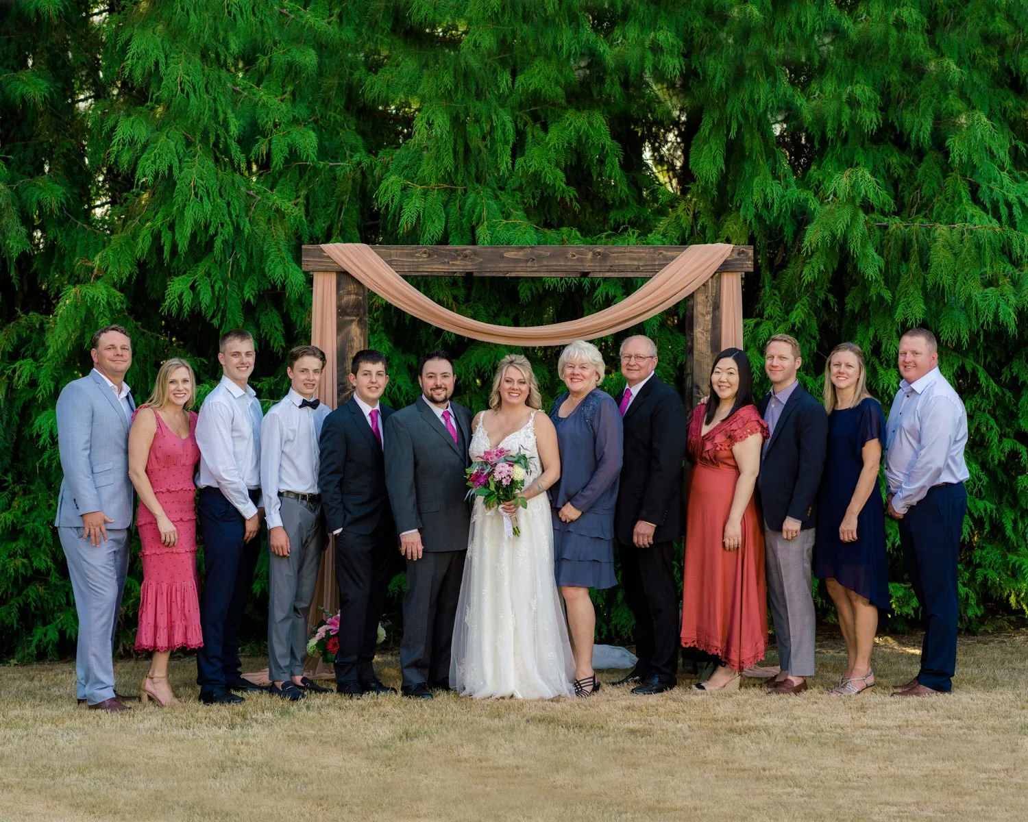 Family portrait at a backyard wedding in Vancouver, Washington with bride and groom standing under a wooden arch draped in blush fabric, surrounded by evergreen trees and wedding guests.