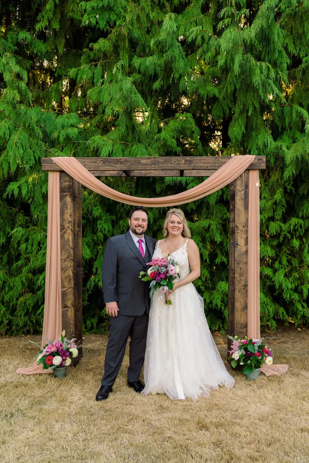 Bride and groom portrait at a Vancouver, WA backyard wedding standing under a rustic wooden ceremony arch with blush draping and colorful floral arrangements against lush green trees.