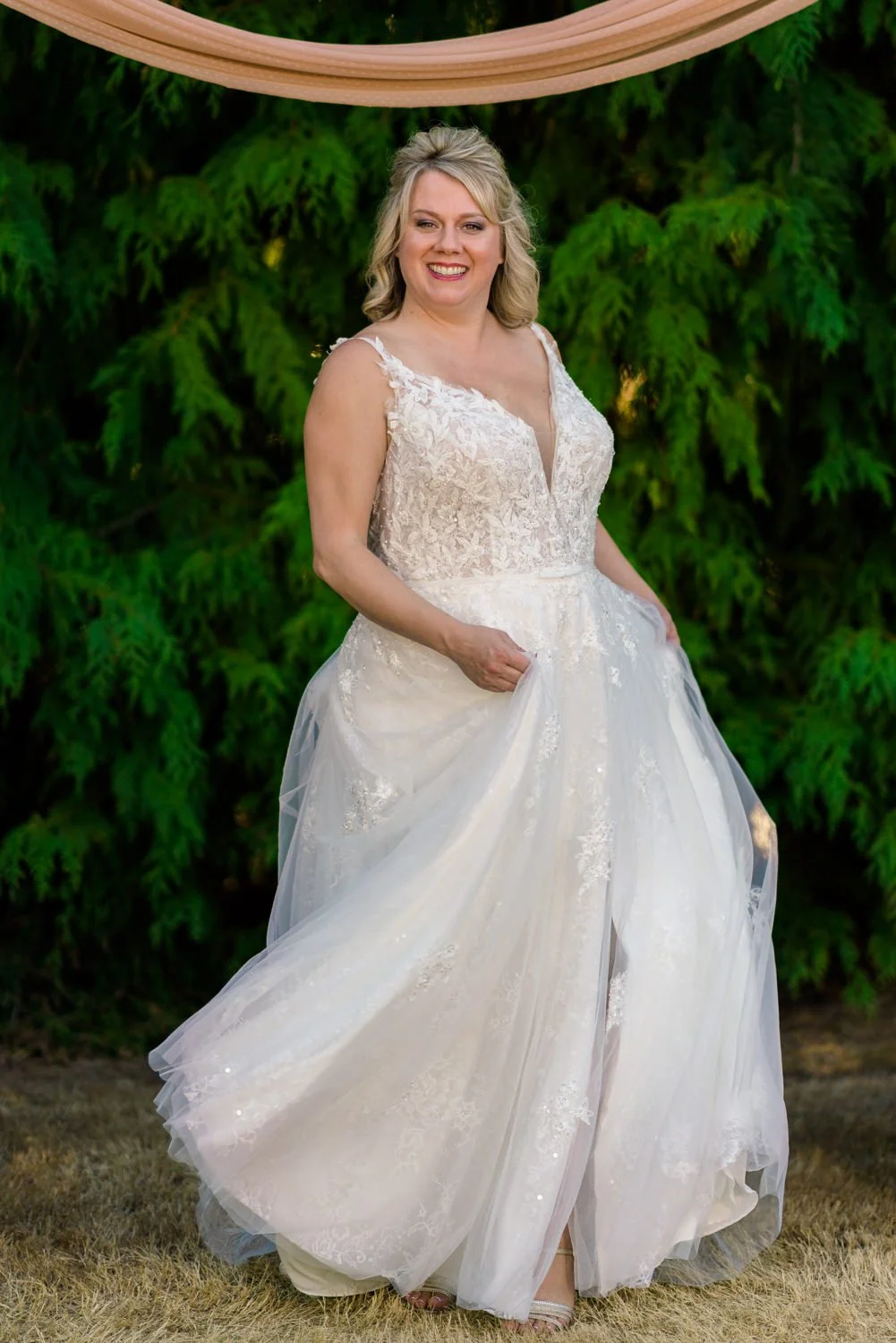Bride portrait at a Vancouver Washington backyard wedding twirling in a lace wedding dress with bouquet in hand, photographed in front of tall evergreen trees during golden hour.